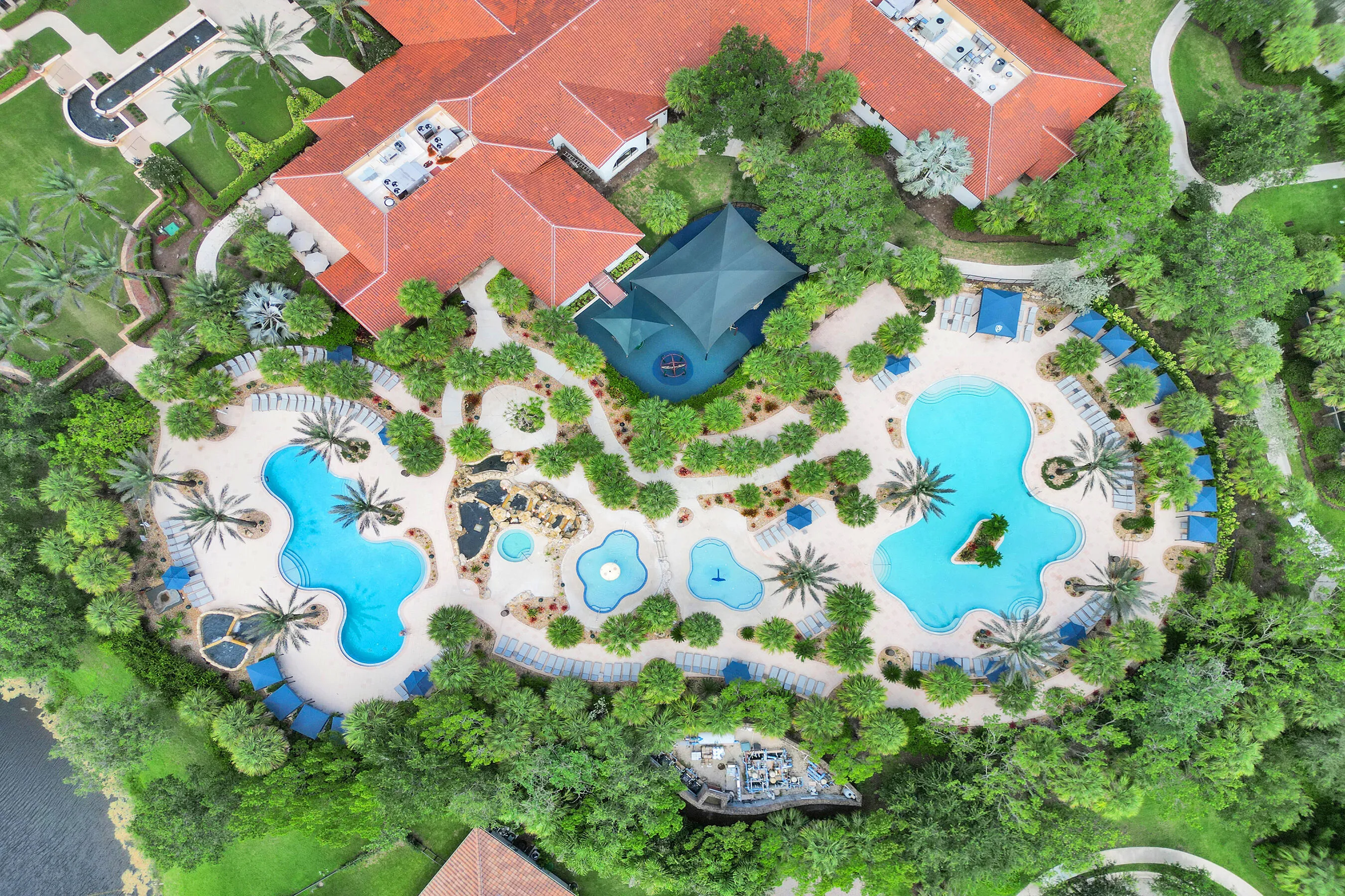 Extensive amenity area featuring multiple swimming pools, mature palm trees, lush landscaping, and a large building with a terracotta roof