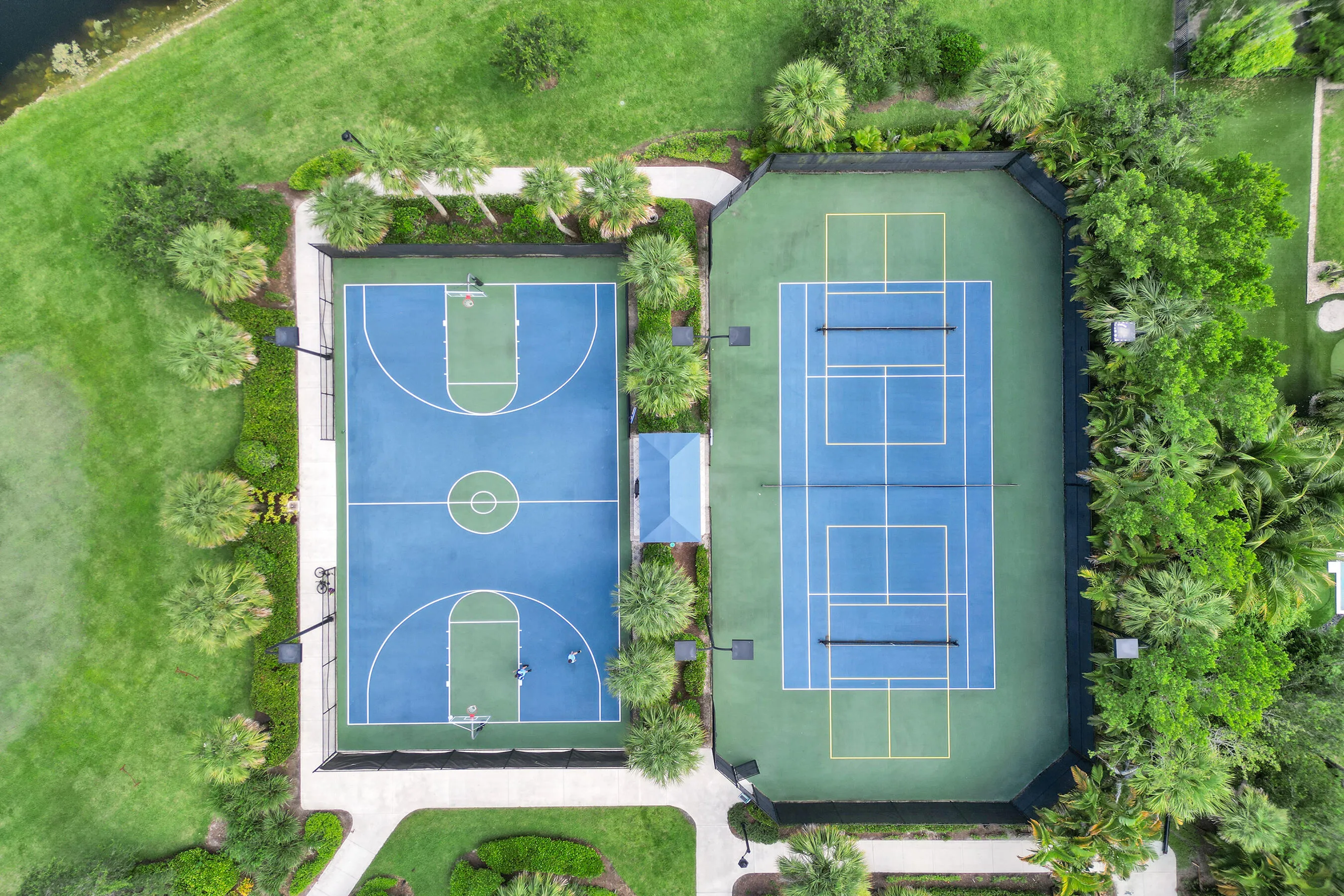 Regulation-size basketball court, two tennis courts, lush surrounding landscaping, paved perimeter pathways, and adjacent manicured green spaces