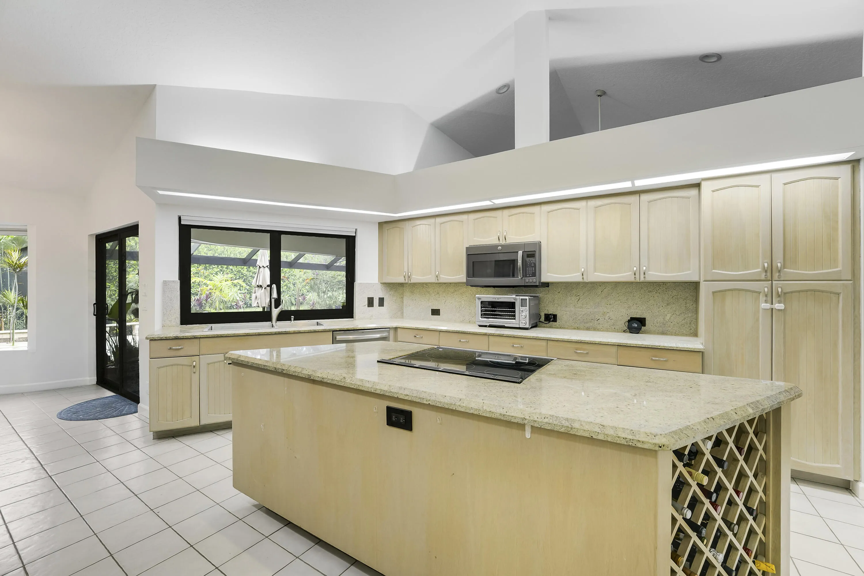Bright, open kitchen featuring a large central island with cooktop and wine rack, stainless appliances, and a wide passthrough window overlooking pool oasis.