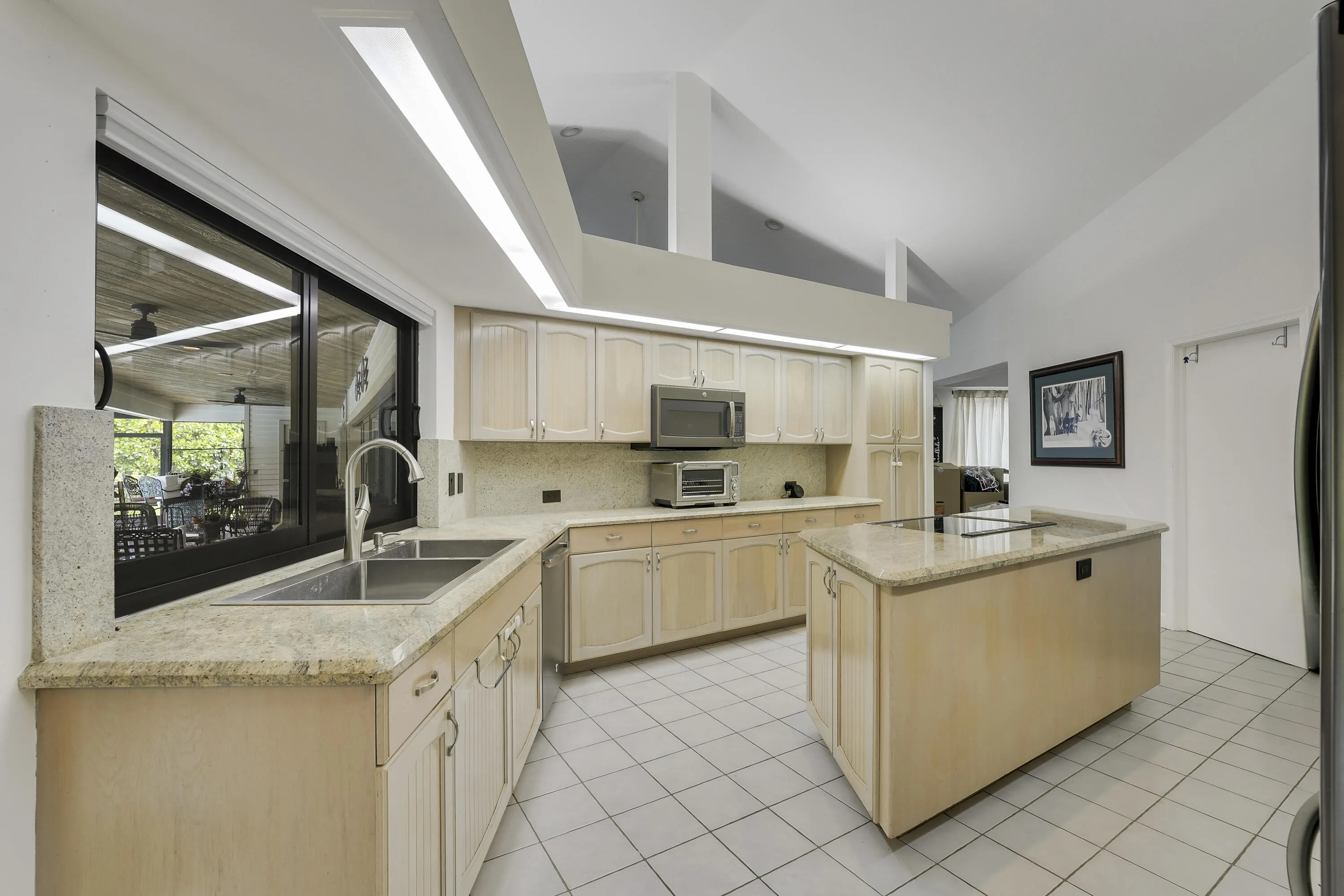 Open kitchen featuring a double sink beneath a passthrough window, spacious island, and architectural ceiling design.