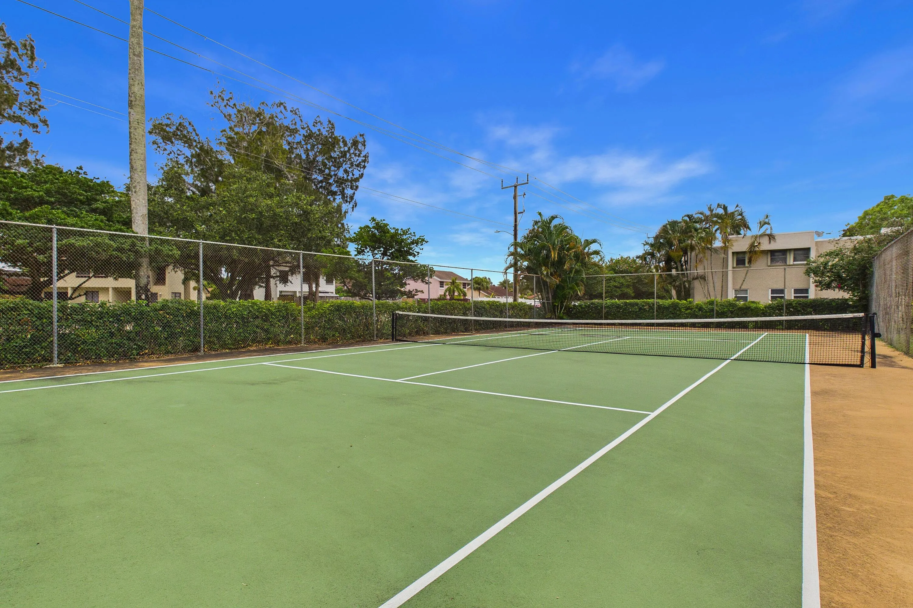 Green hard tennis court with white line markings, surrounded by a chain-link fence and mature landscaping