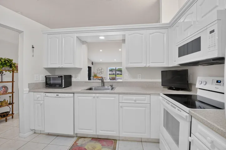 Bright, open kitchen featuring white cabinetry with raised-panel doors, stainless steel appliances including double wall ovens, granite countertops, undermount sink with chrome fixtures, and tile flooring
