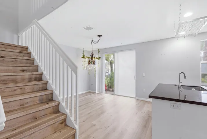 Wood-finish staircase with white balusters, leading to a spacious living area featuring wood-finish flooring, a contemporary chandelier, and a sliding glass door