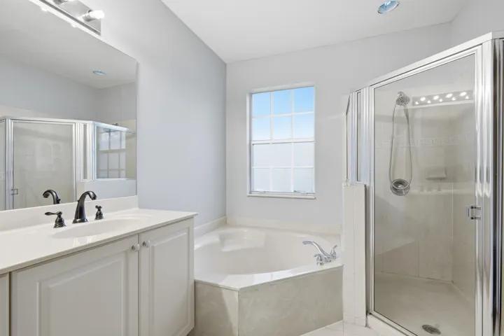 White vanity with a single basin sink and dark-finish fixtures, a built-in corner bathtub, a glass-enclosed shower, and a large window