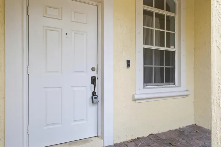 Textured exterior finish, white paneled entry door, double-hung window with grid, decorative window trim, brick paver flooring