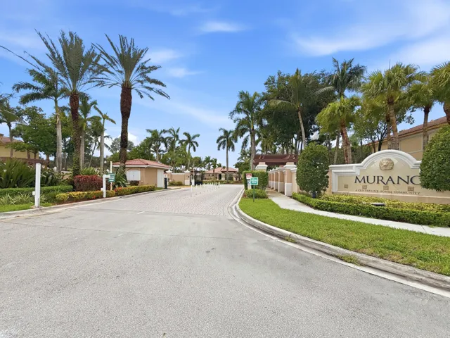 Entryway featuring a decorative sign, lush landscaping, mature palm trees, paved road, and a guardhouse structure