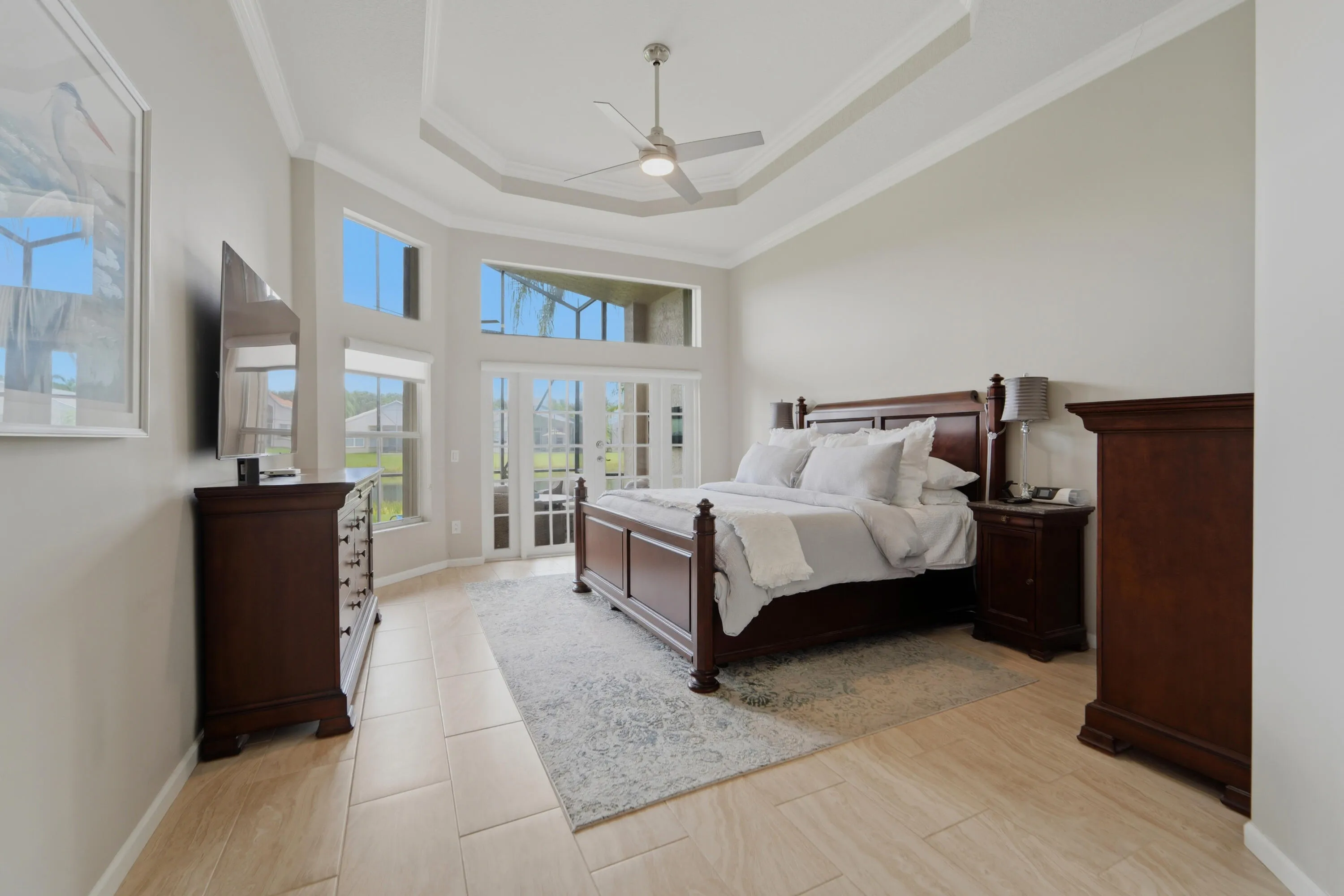 Spacious primary suite with en-suite bath and two walk in closets features a tray ceiling, natural light and French doors opening to the covered lanai.