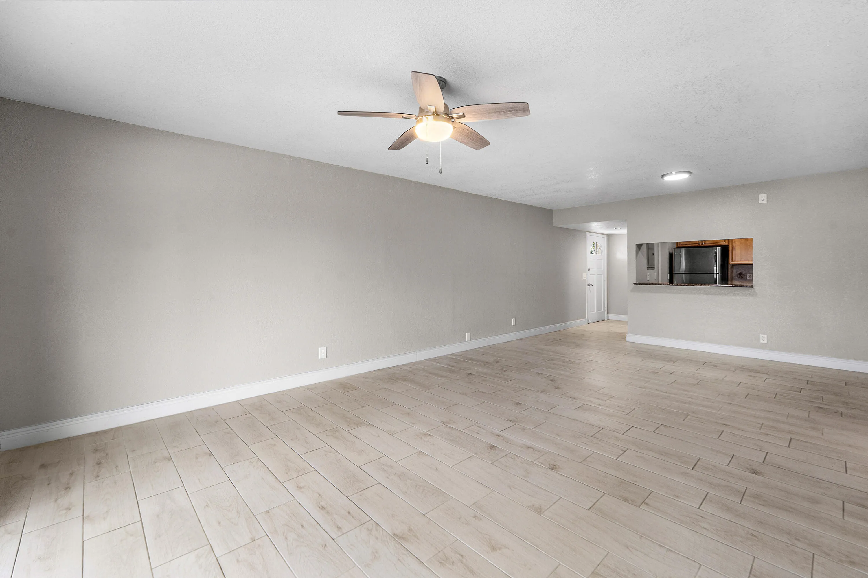 Spacious living area featuring light-toned tile flooring, neutral wall tones, a ceiling fan with integrated lighting, and a pass-through window to the kitchen