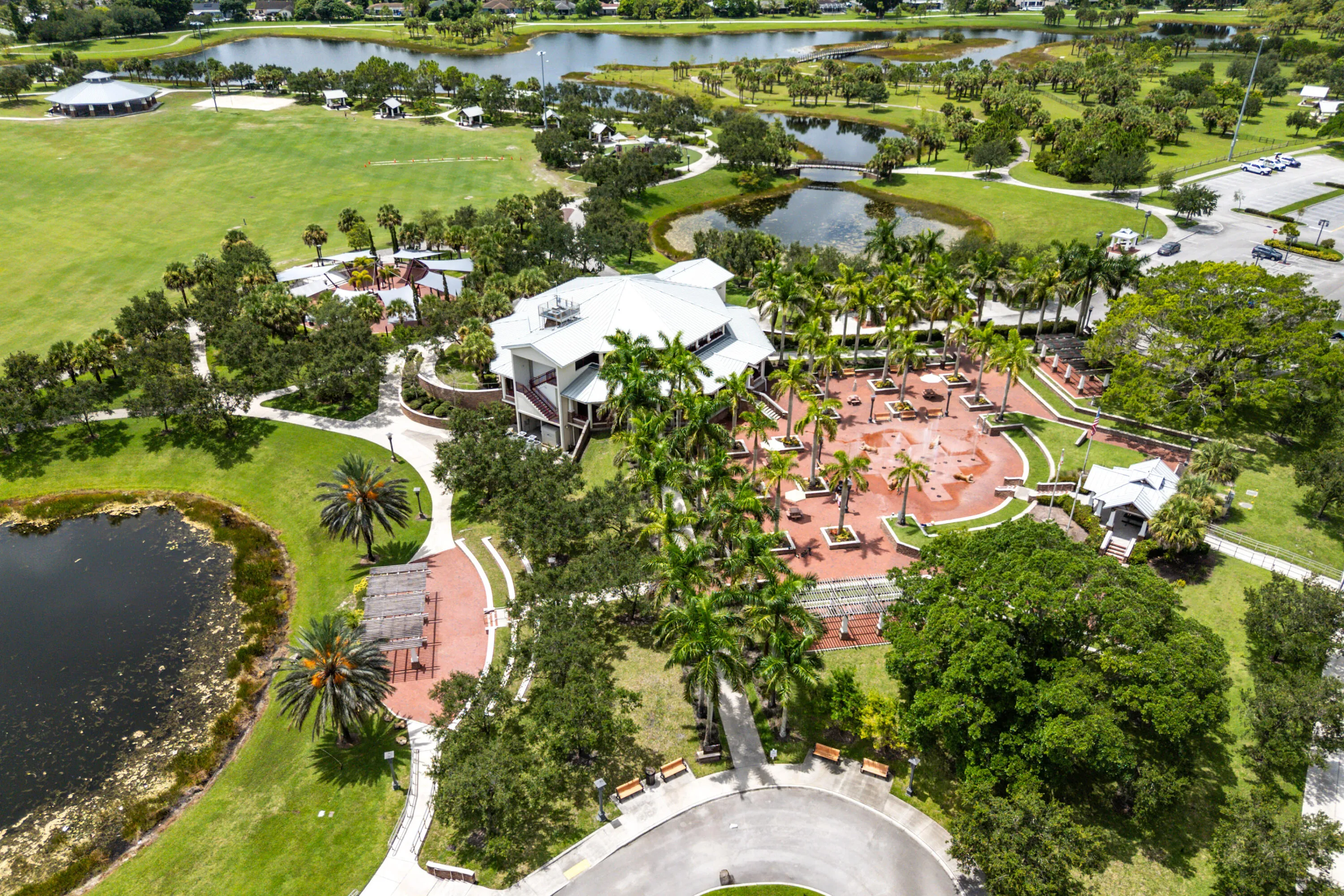 Expansive park grounds featuring a large lake, mature trees, open green spaces, paved walking paths, and a prominent multi-story building with a white metal roof