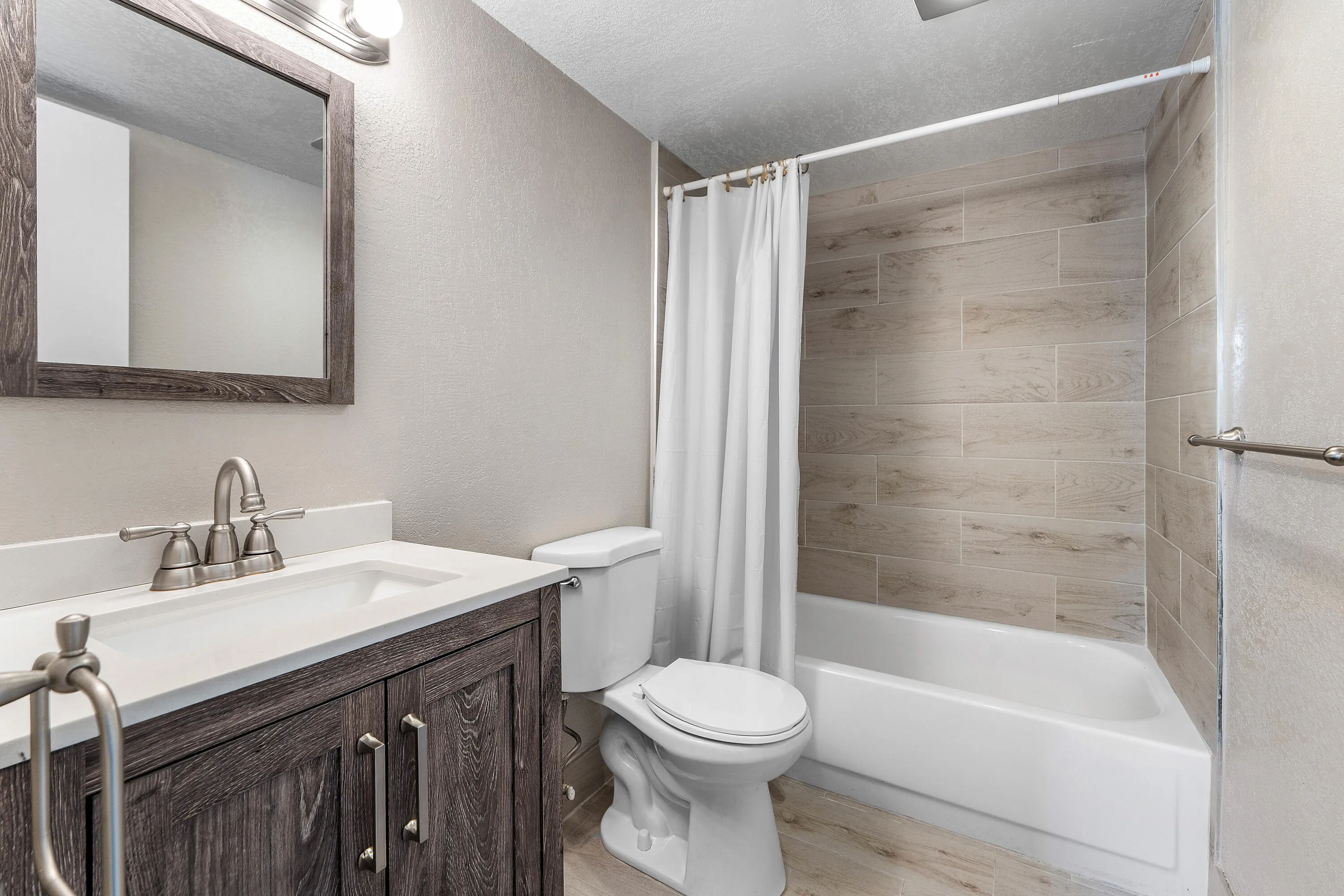 Wood-finish vanity with a white countertop and integrated sink, framed mirror, and a tub-shower combination with wood-look tiled walls