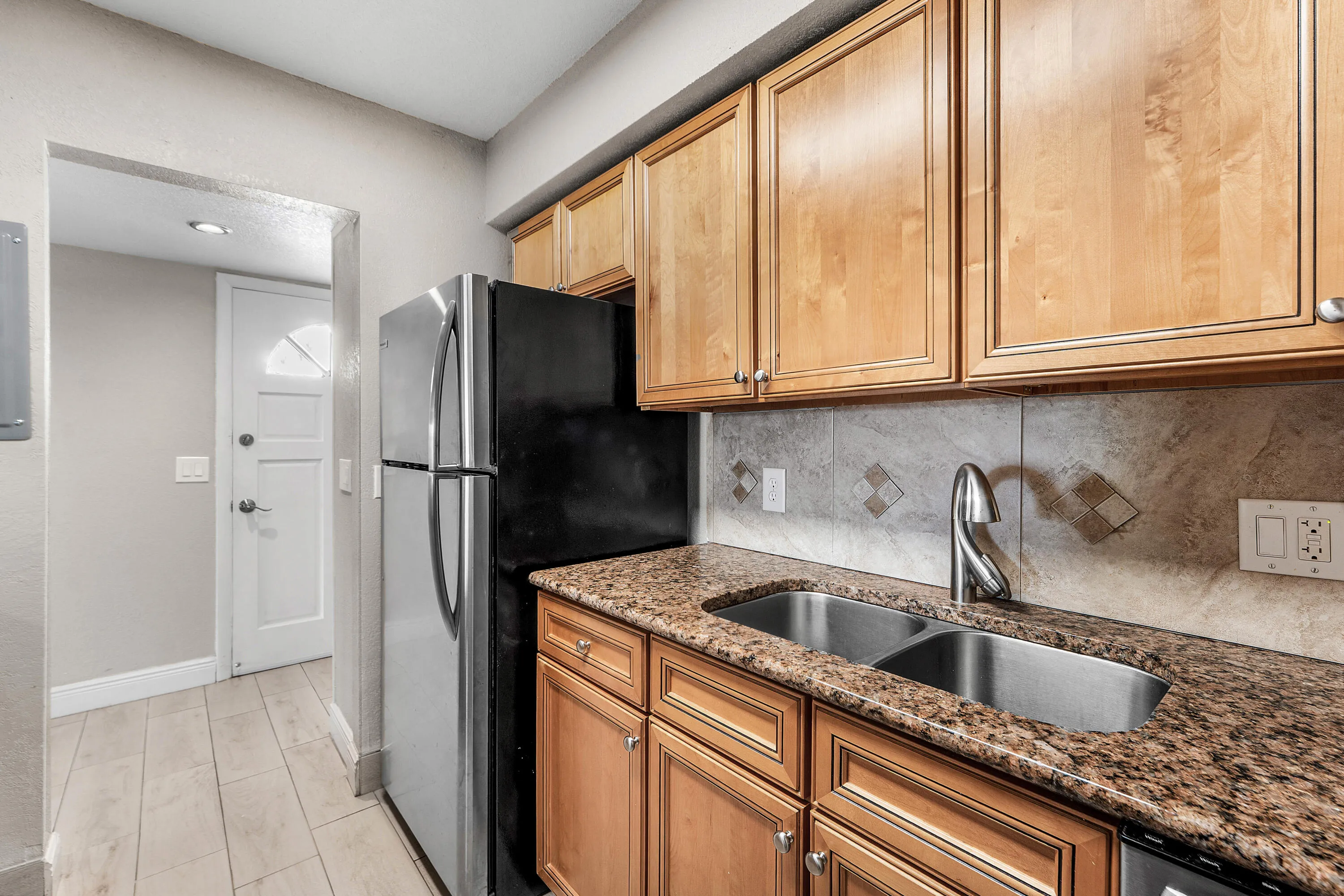 Wood-finish cabinetry, stone-look countertops, stainless steel sink, and tile backsplash with decorative accents