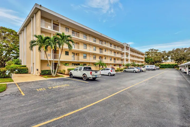 Multi-story building featuring balconies with white railings, a paved parking area, and manicured landscaping