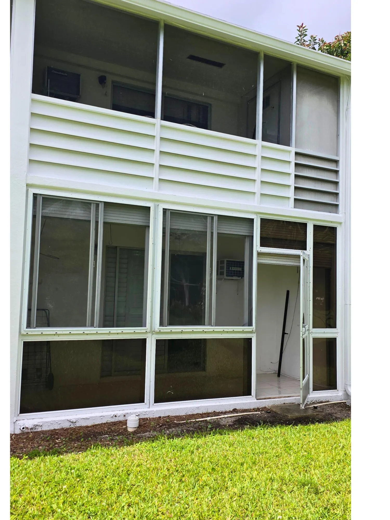Modern two-story residence featuring expansive glazing with aluminum-frame windows and glass sliding doors across the lower level, horizontal louvered privacy screening on the upper deck, and manicured lawn with mature landscaping