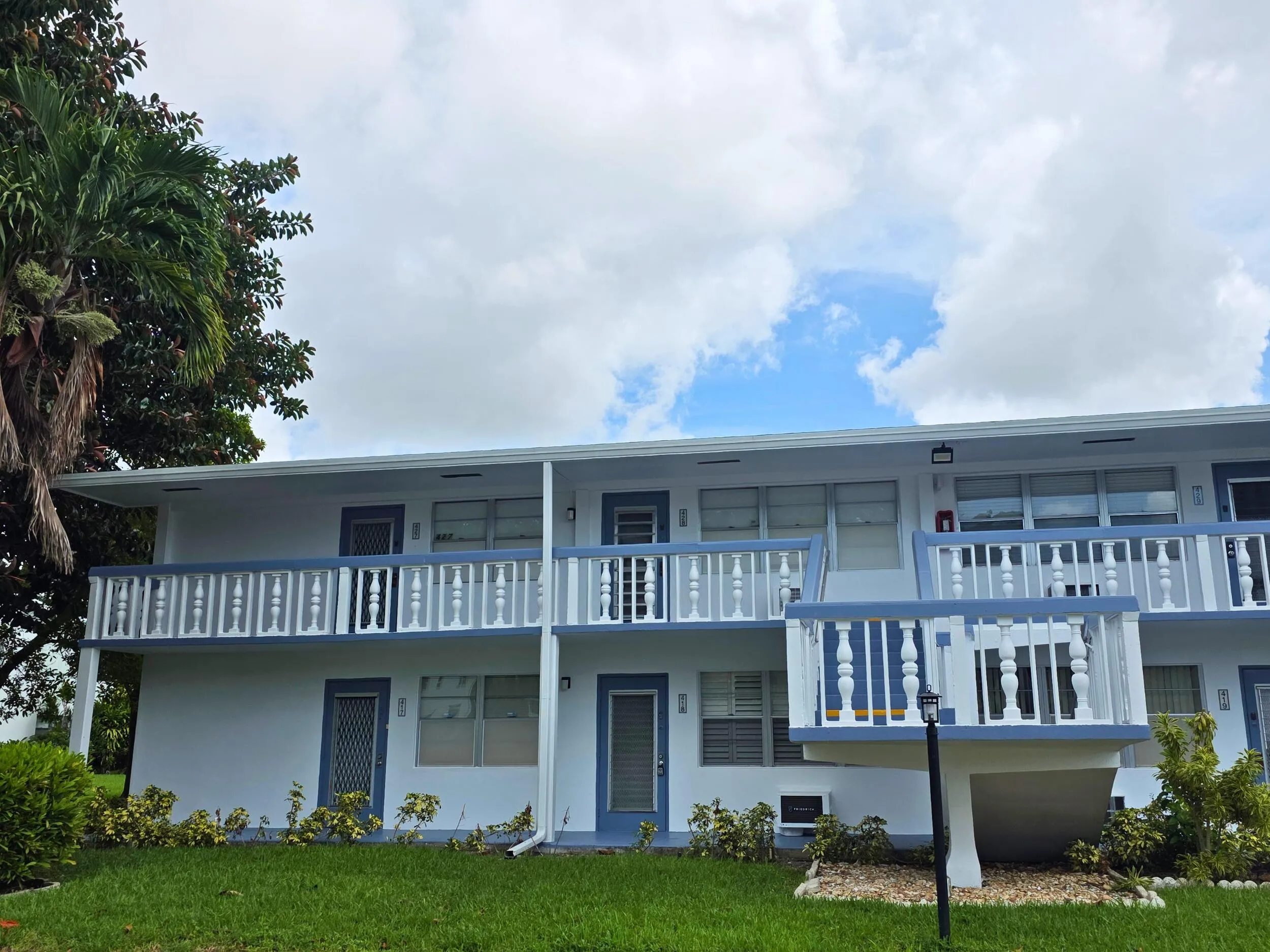 Two-story building featuring white exterior, blue trim accents, multiple balconies with white and blue balusters, and a green lawn with landscaping