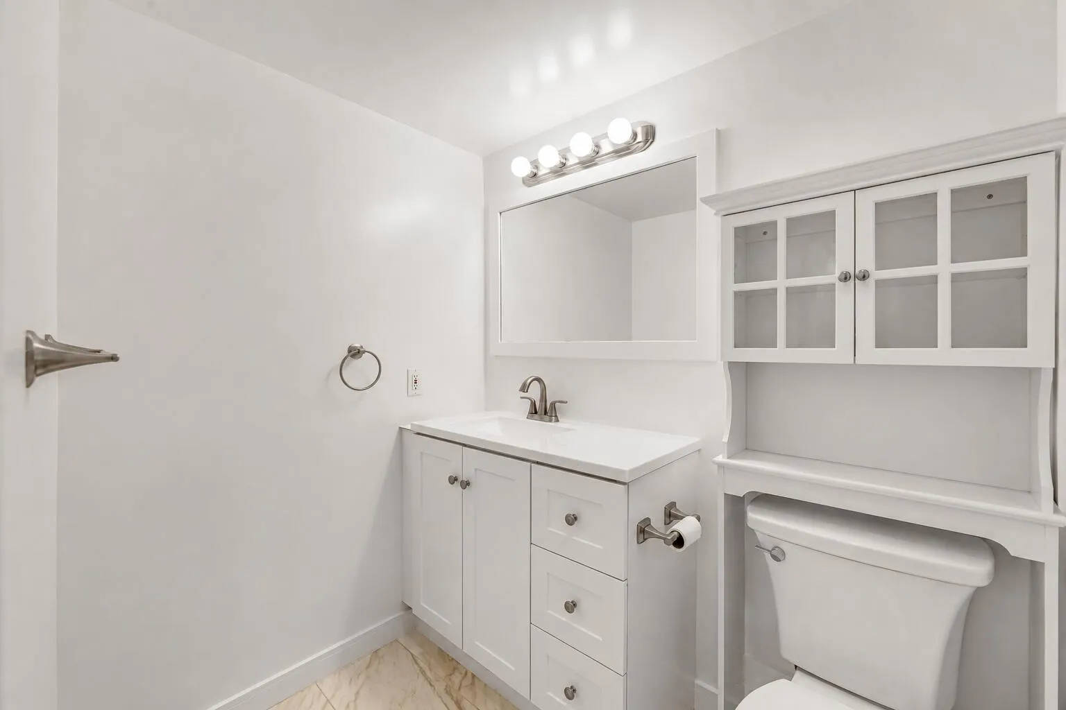 Bright bathroom featuring white cabinetry with multiple drawers and hardware accents, a white vanity with an undermount sink and brushed nickel faucet, framed mirror with integrated lighting, and white glass-front wall cabinet with shelving