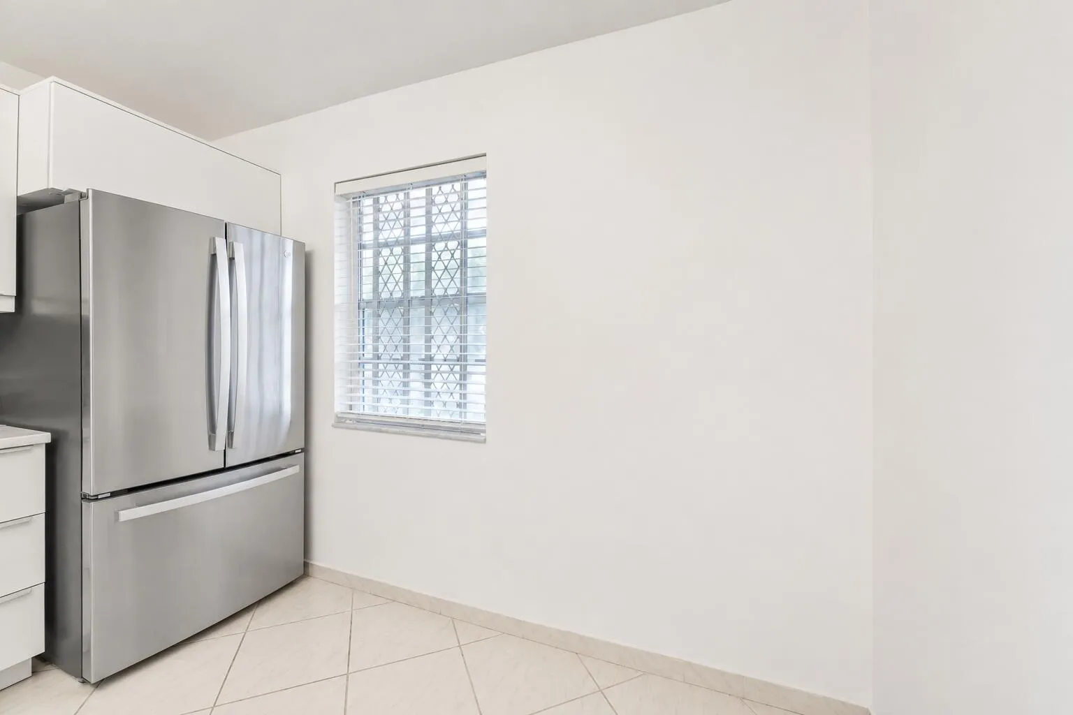 Stainless steel French door refrigerator, white upper cabinetry, tiled flooring, and a single window with blinds and security bars