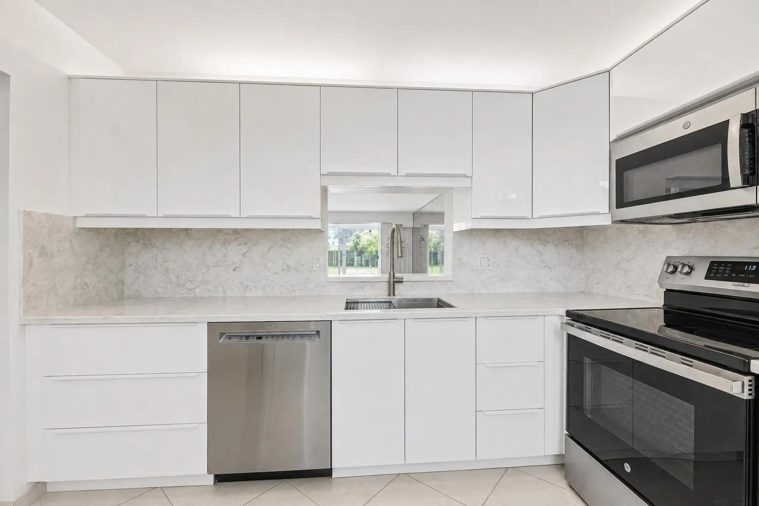 Modern kitchen featuring white cabinetry with clean-lined hardware, marble-look backsplash, stainless steel appliances including a built-in microwave and double wall ovens, and a dedicated dishwasher with a center window overlooking natural light