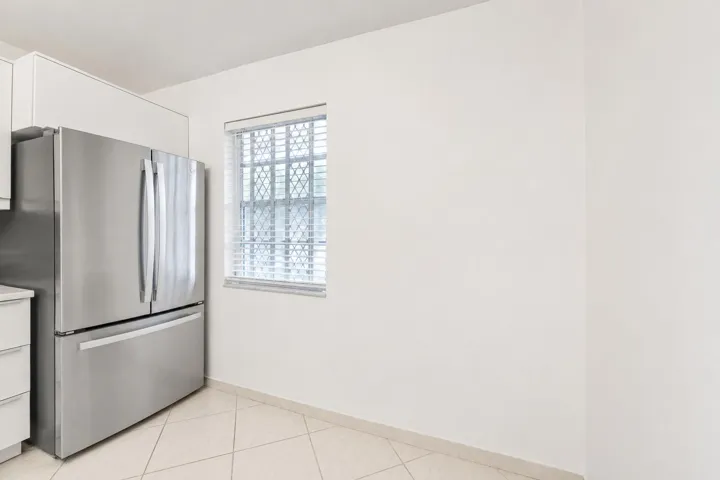 Stainless steel French door refrigerator, white upper cabinetry, tiled flooring, and a single window with blinds and security bars