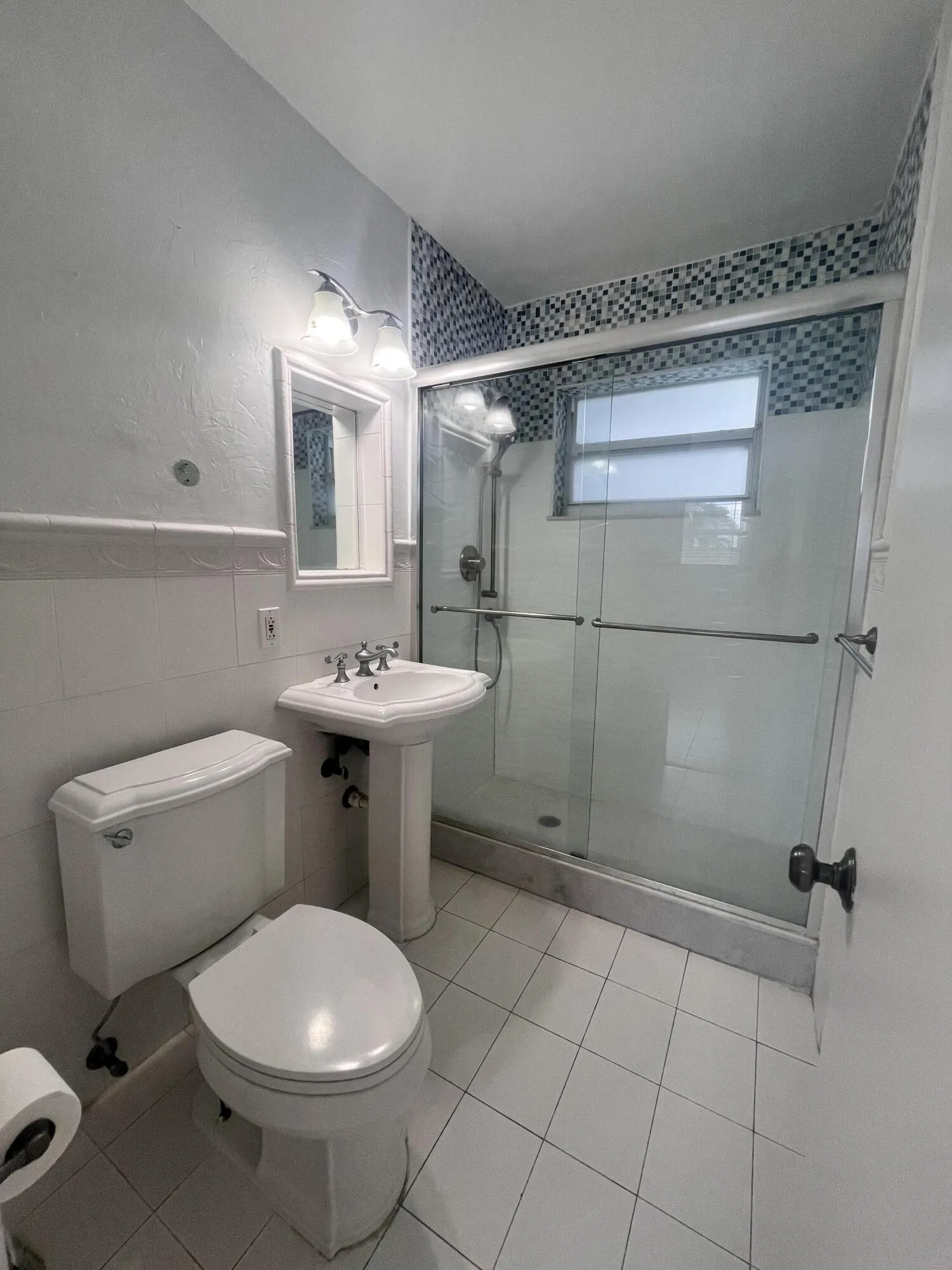 Pedestal sink with chrome fixtures, tiled shower with glass enclosure, mosaic accent tile, and white floor tiling