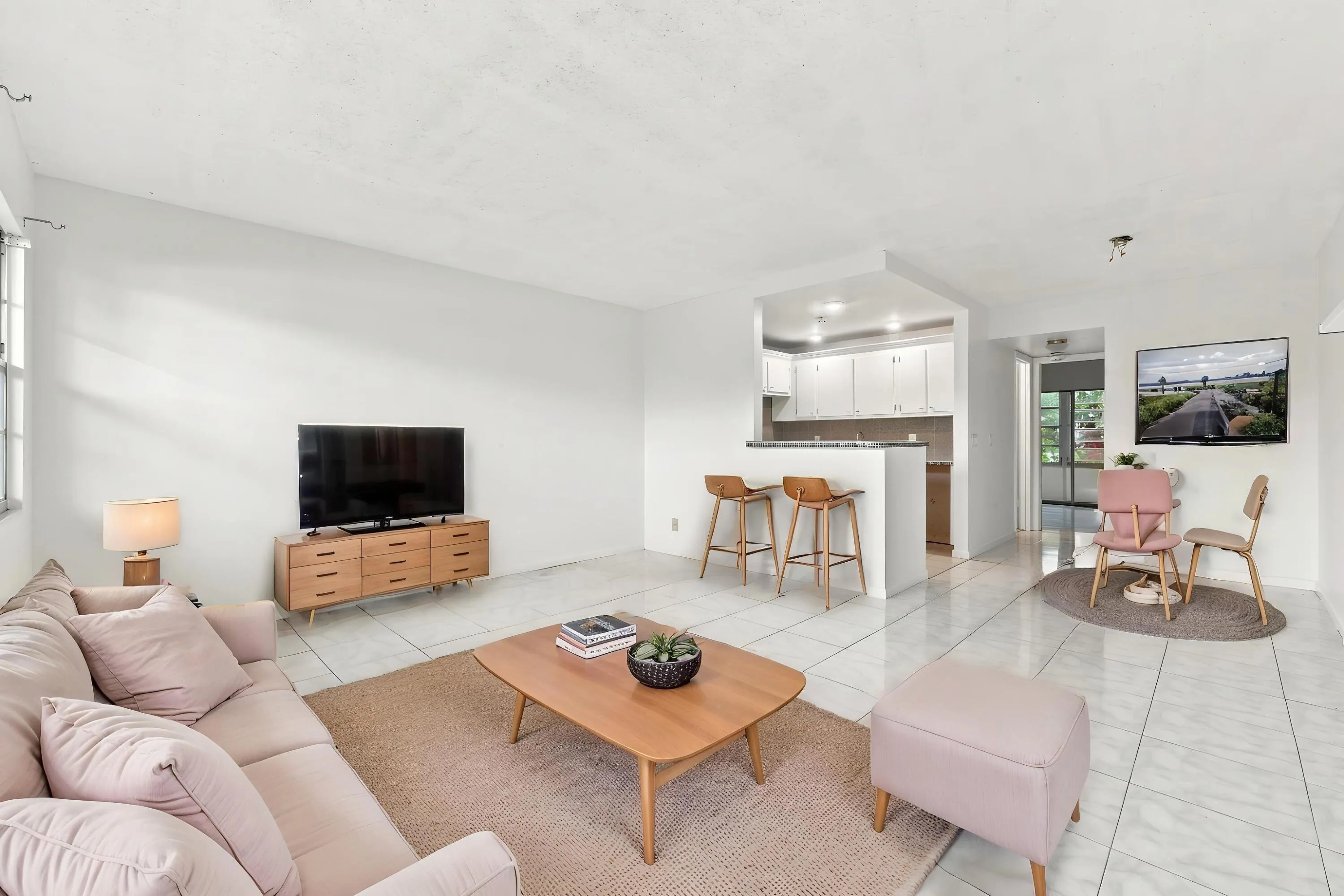 Spacious and bright living area featuring tile flooring throughout, abundant natural light, and a large wall of windows overlooking lush tropical greenery. The open layout flows seamlessly into the kitchen and offers plenty of flexibility for comfortable everyday living and entertaining.