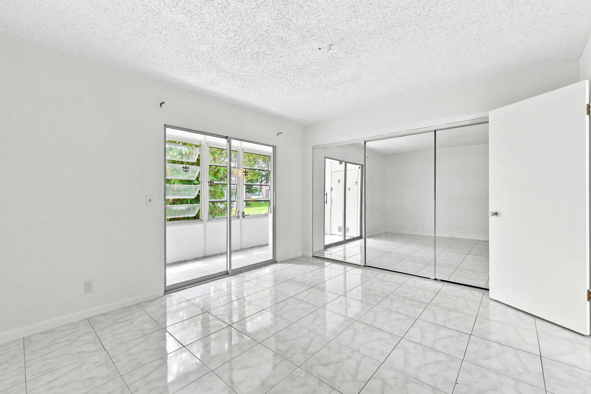 Spacious bedroom featuring tile flooring, an expansive mirrored closet for generous storage, and sliding glass doors opening directly to the enclosed patio. Abundant natural light and lush tropical views create a bright, inviting retreat.