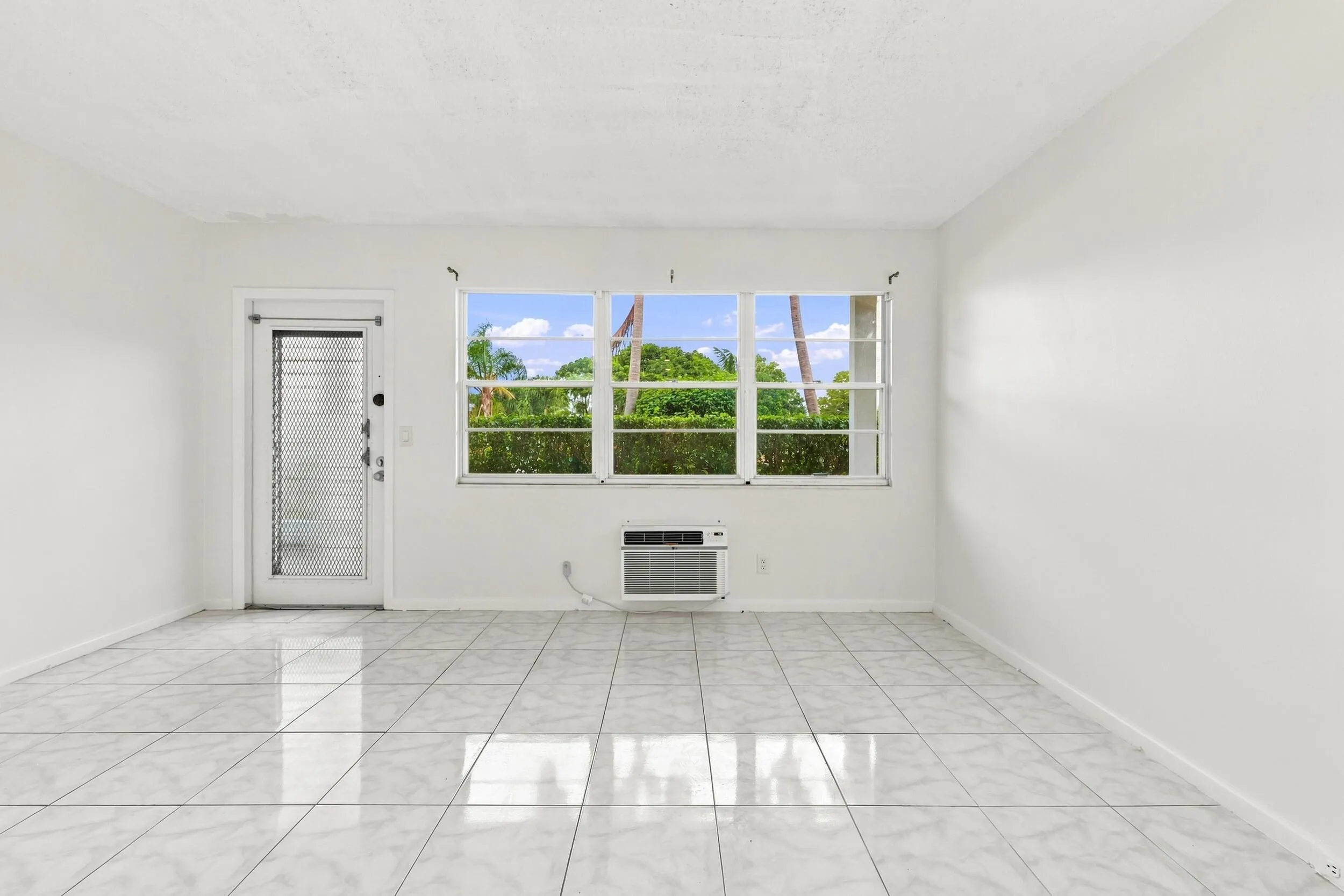 Spacious and bright living area featuring tile flooring throughout, abundant natural light, and a large wall of windows overlooking lush tropical greenery. The open layout flows seamlessly into the kitchen and offers plenty of flexibility for comfortable everyday living and entertaining.