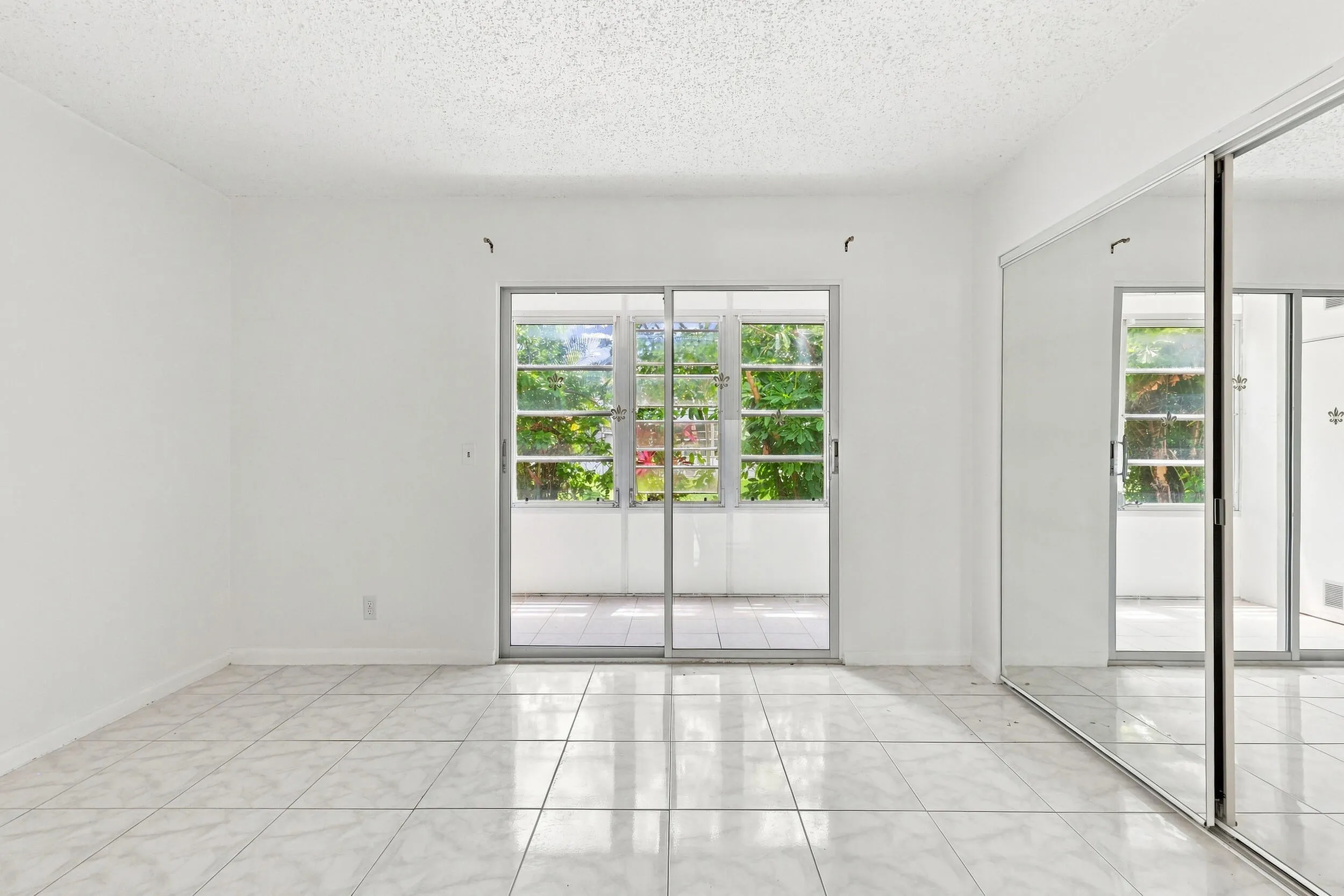 Spacious bedroom featuring tile flooring, an expansive mirrored closet for generous storage, and sliding glass doors opening directly to the enclosed patio. Abundant natural light and lush tropical views create a bright, inviting retreat.