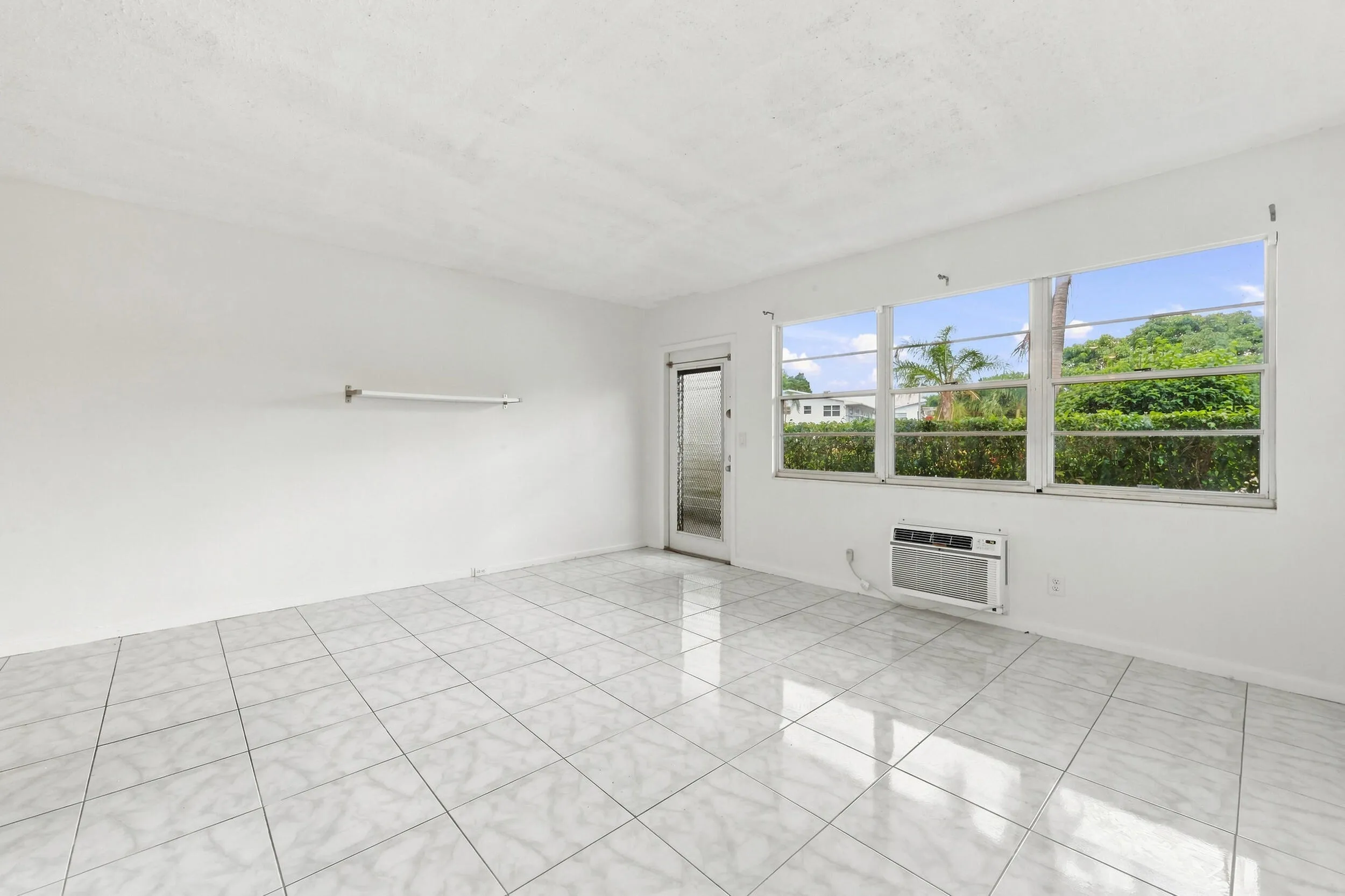 Spacious and bright living area featuring tile flooring throughout, abundant natural light, and a large wall of windows overlooking lush tropical greenery. The open layout flows seamlessly into the kitchen and offers plenty of flexibility for comfortable everyday living and entertaining.