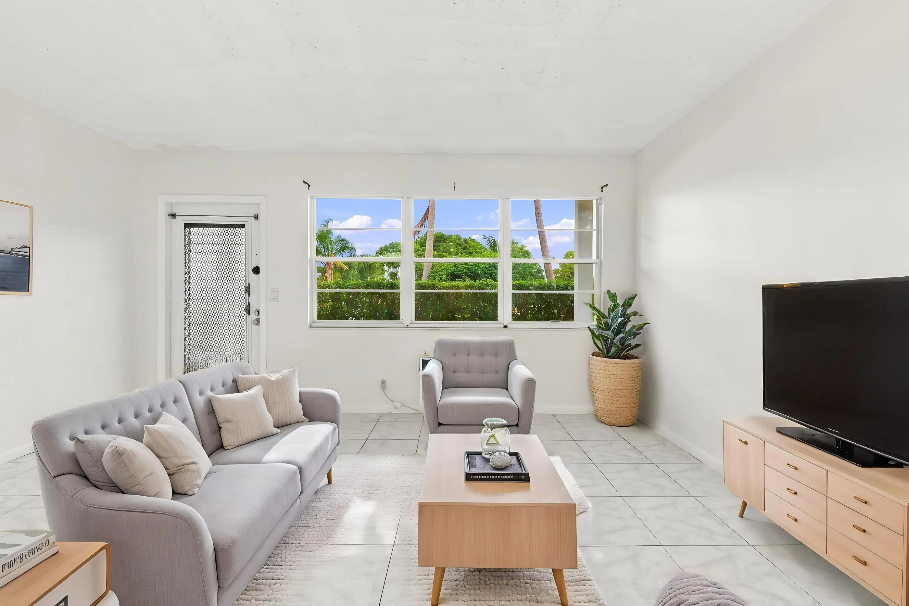 Spacious and bright living area featuring tile flooring throughout, abundant natural light, and a large wall of windows overlooking lush tropical greenery. The open layout flows seamlessly into the kitchen and offers plenty of flexibility for comfortable everyday living and entertaining.