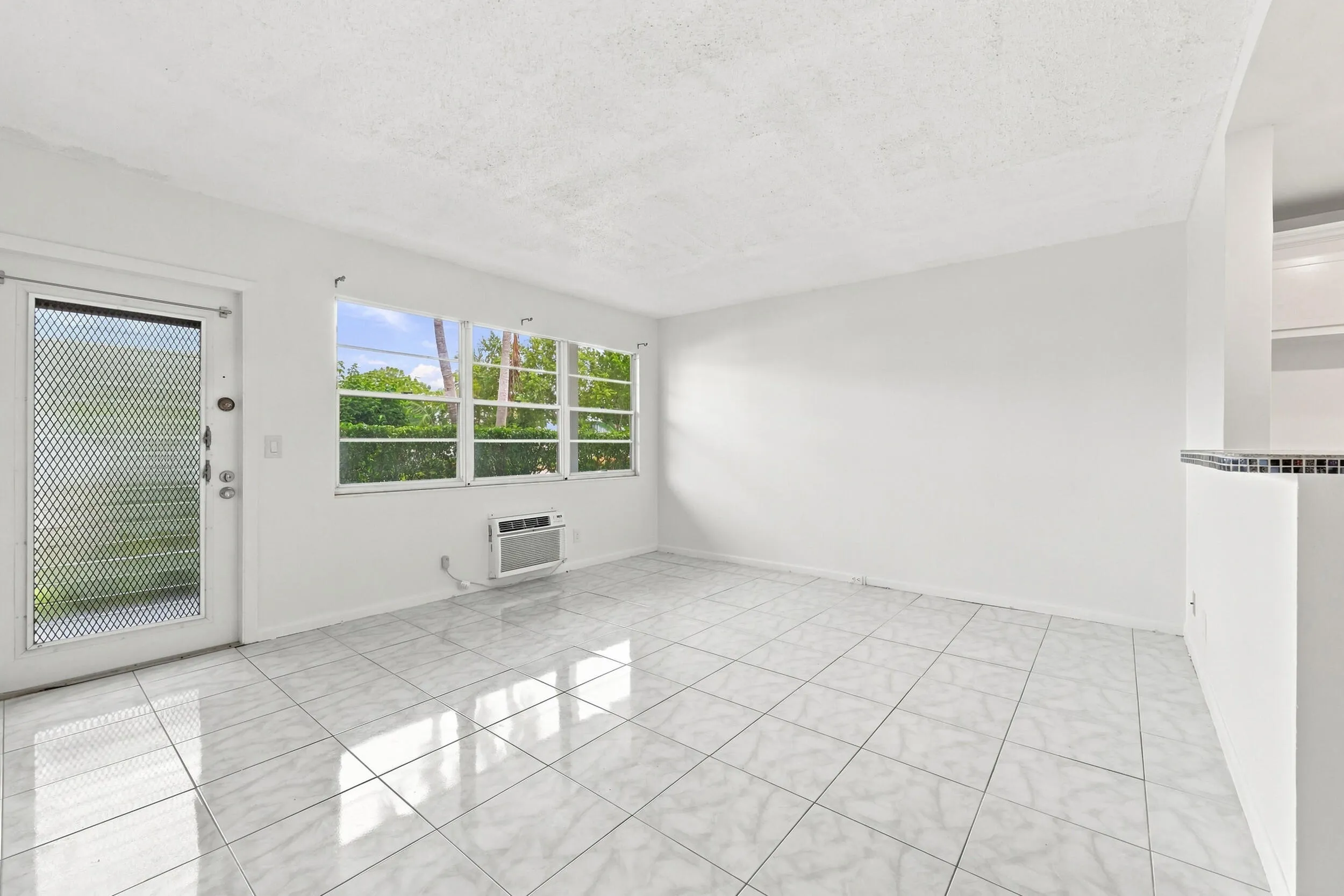 Spacious and bright living area featuring tile flooring throughout, abundant natural light, and a large wall of windows overlooking lush tropical greenery. The open layout flows seamlessly into the kitchen and offers plenty of flexibility for comfortable everyday living and entertaining.