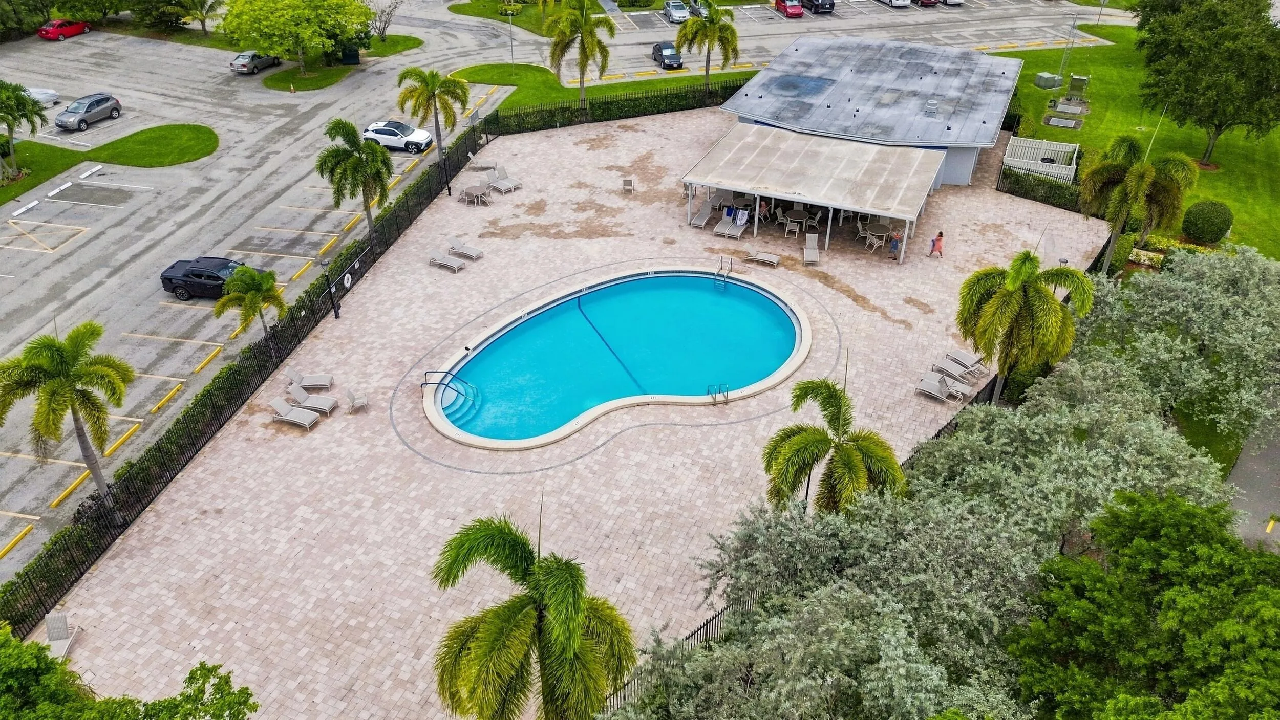 Aerial views showcase the expansive community pool and spacious surrounding sundeck, complete with abundant lounge areas, a covered seating pavilion, tropical landscaping, and plenty of space to relax and enjoy the South Florida sunshine.
