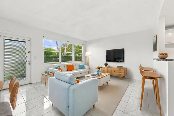 Spacious and bright living area featuring tile flooring throughout, abundant natural light, and a large wall of windows overlooking lush tropical greenery. The open layout flows seamlessly into the kitchen and offers plenty of flexibility for comfortable everyday living and entertaining.
