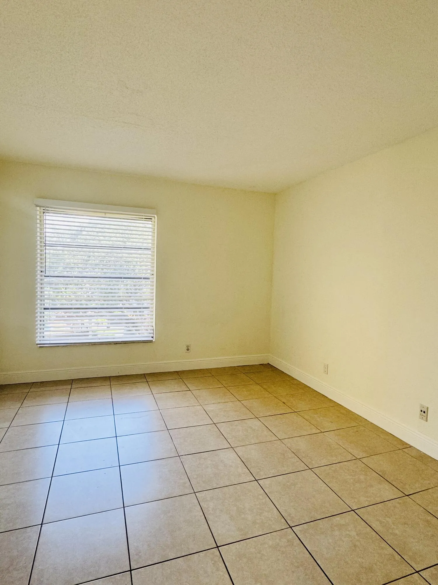 Bright, sunlit bedroom featuring ceramic tile flooring, large window with horizontal blinds providing natural light, and neutral yellow walls throughout the space