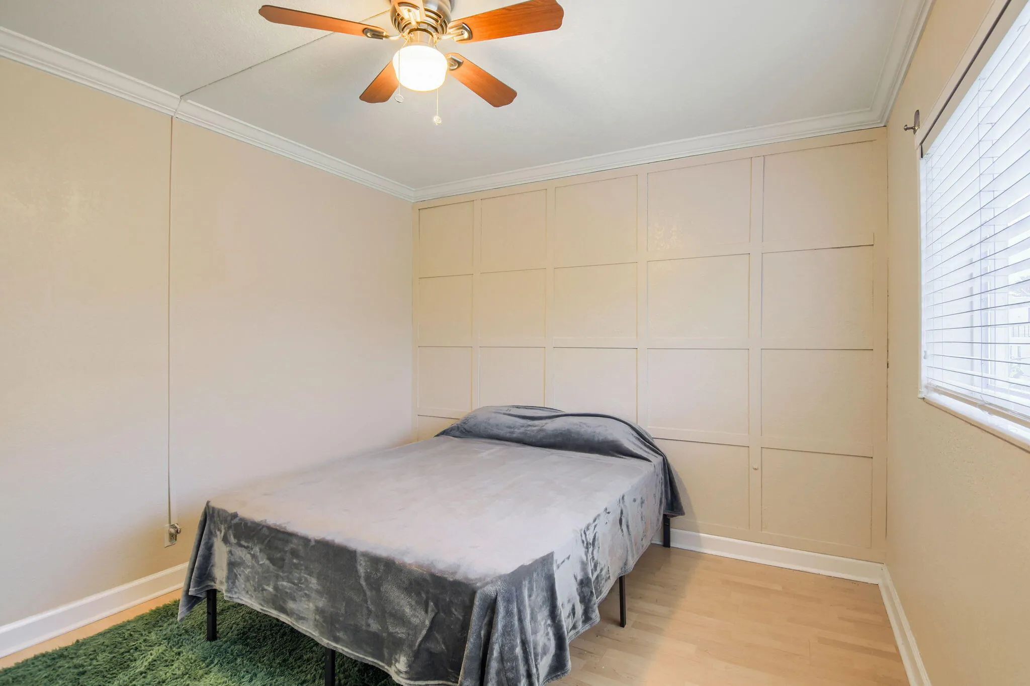 Wood-finish flooring with crown molding, a decorative accent wall, a window with blinds, and a ceiling fan with integrated lighting