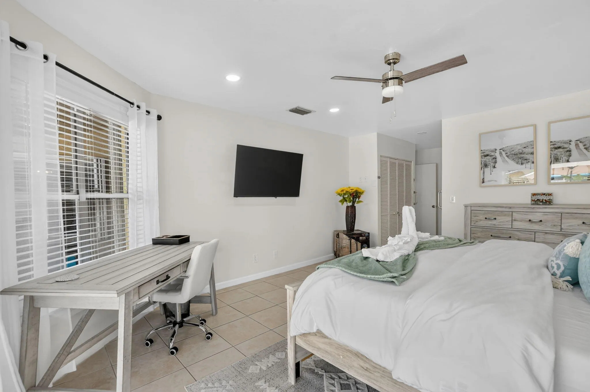 Spacious primary bedroom featuring tile flooring, a ceiling fan with integrated lighting, a wall-mounted flat-screen television, and a dedicated work desk with office chair