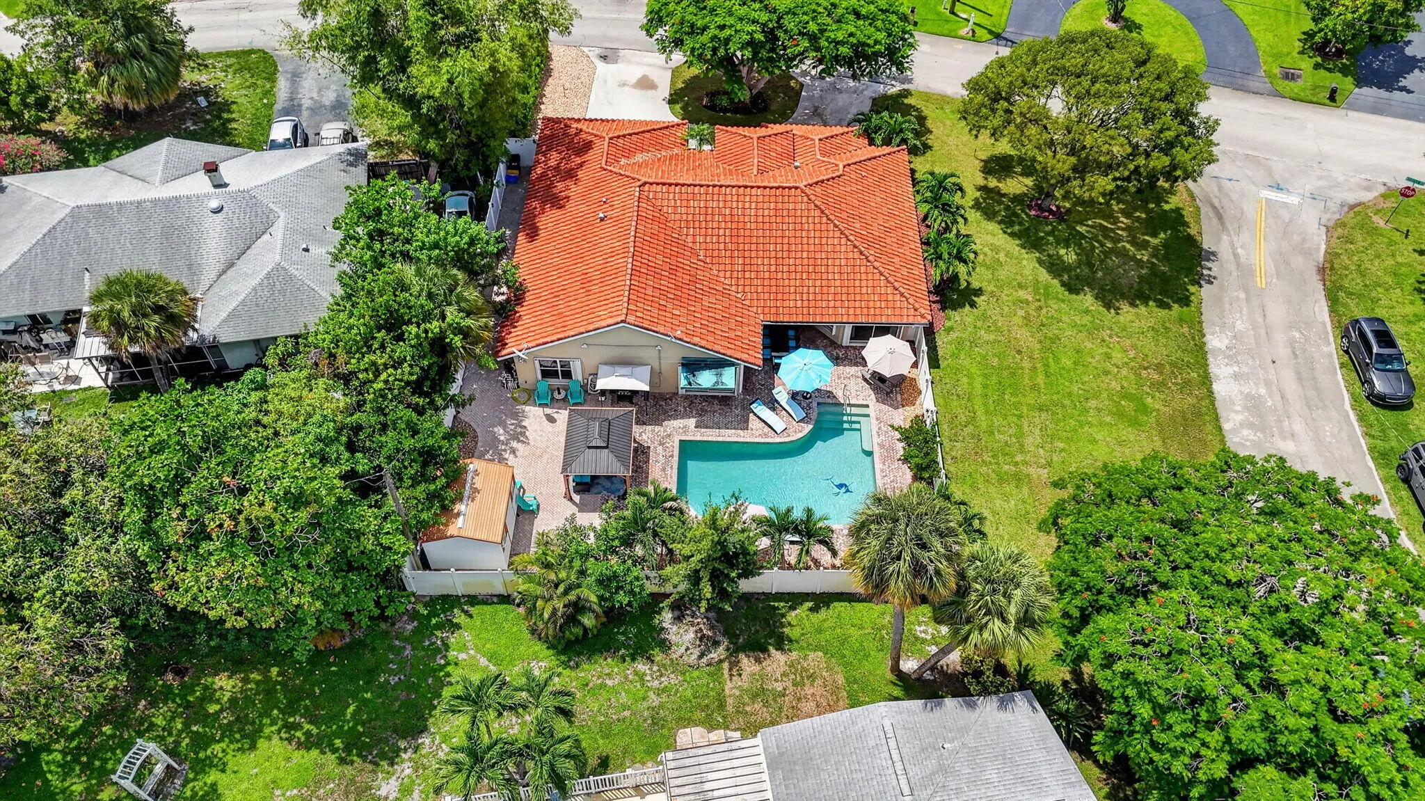 Aerial view of a Spanish Colonial residence featuring a distinctive barrel tile roof, resort-style saltwater pool with surrounding hardscape and lounge areas, mature landscaping with palm trees and lush greenery, and manicured lawn grounds situated on an expansive lot