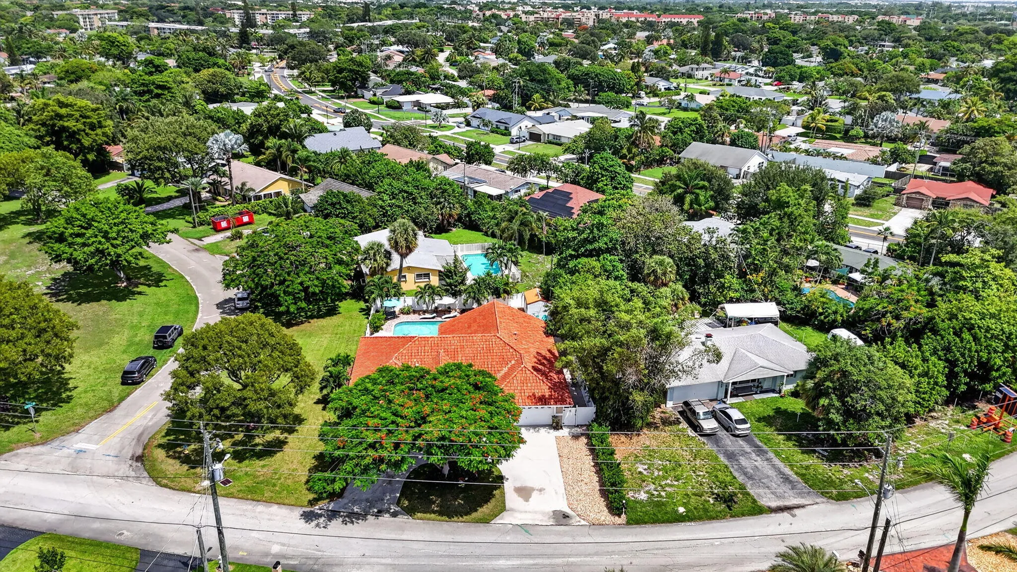 Aerial view showcasing a substantial corner-lot property with a clay tile roof, mature landscaping throughout the grounds, a sparkling swimming pool with surrounding hardscape, and lush tropical vegetation providing privacy and shade across the expansive grounds