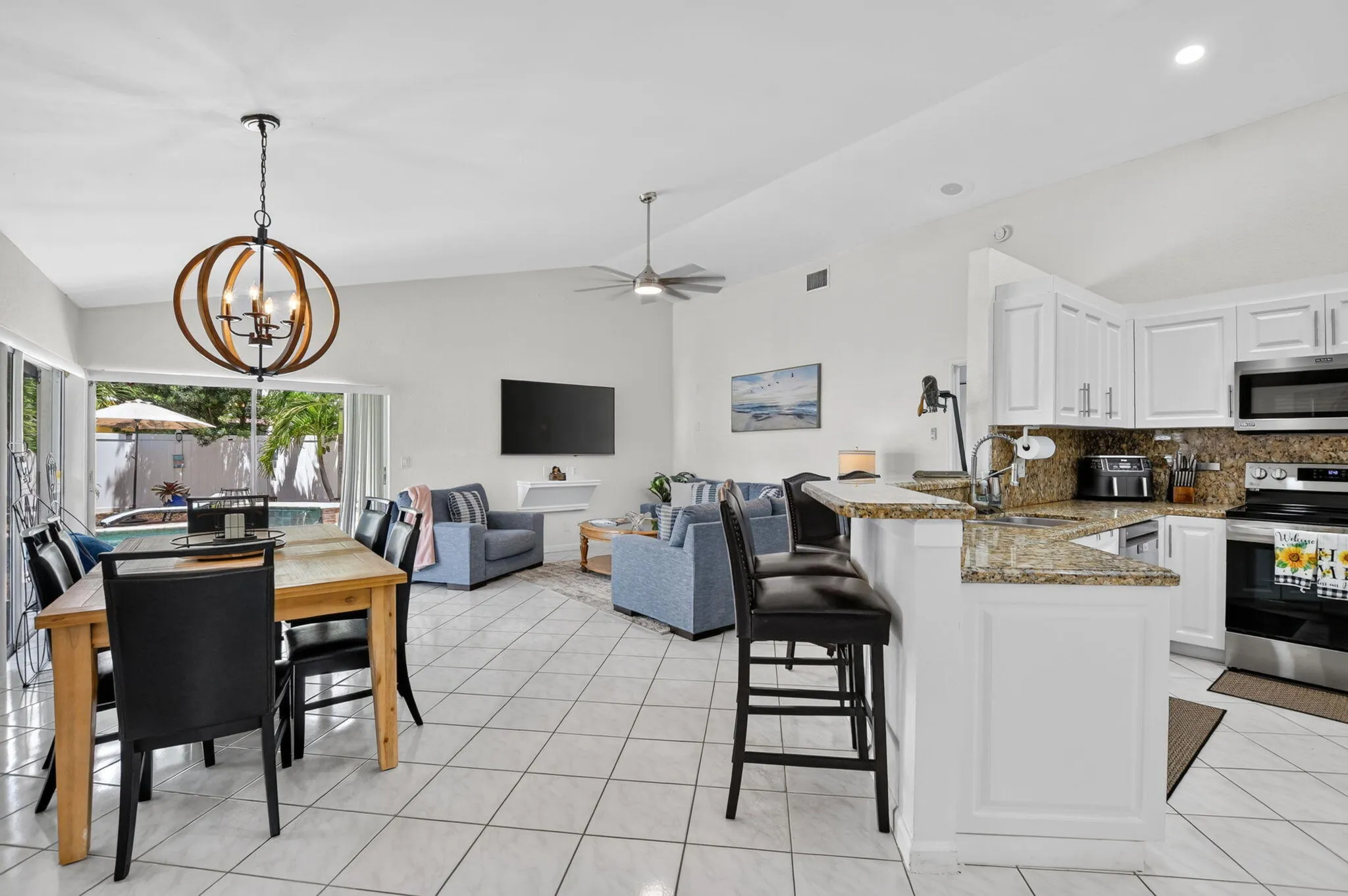 Open-concept living and kitchen area featuring light-filled spaces with large glass sliding doors, white cabinetry with granite countertops, stainless steel appliances, tile flooring throughout, a decorative pendant light fixture, and a ceiling fan