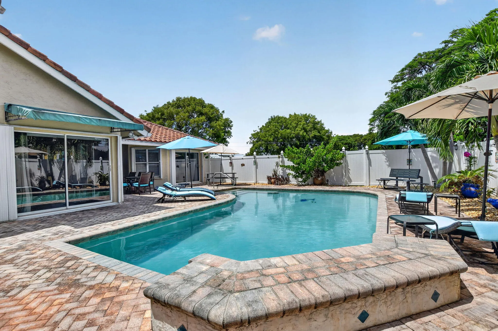 Outdoor swimming pool with a brick paver deck, white privacy fence, and mature landscaping