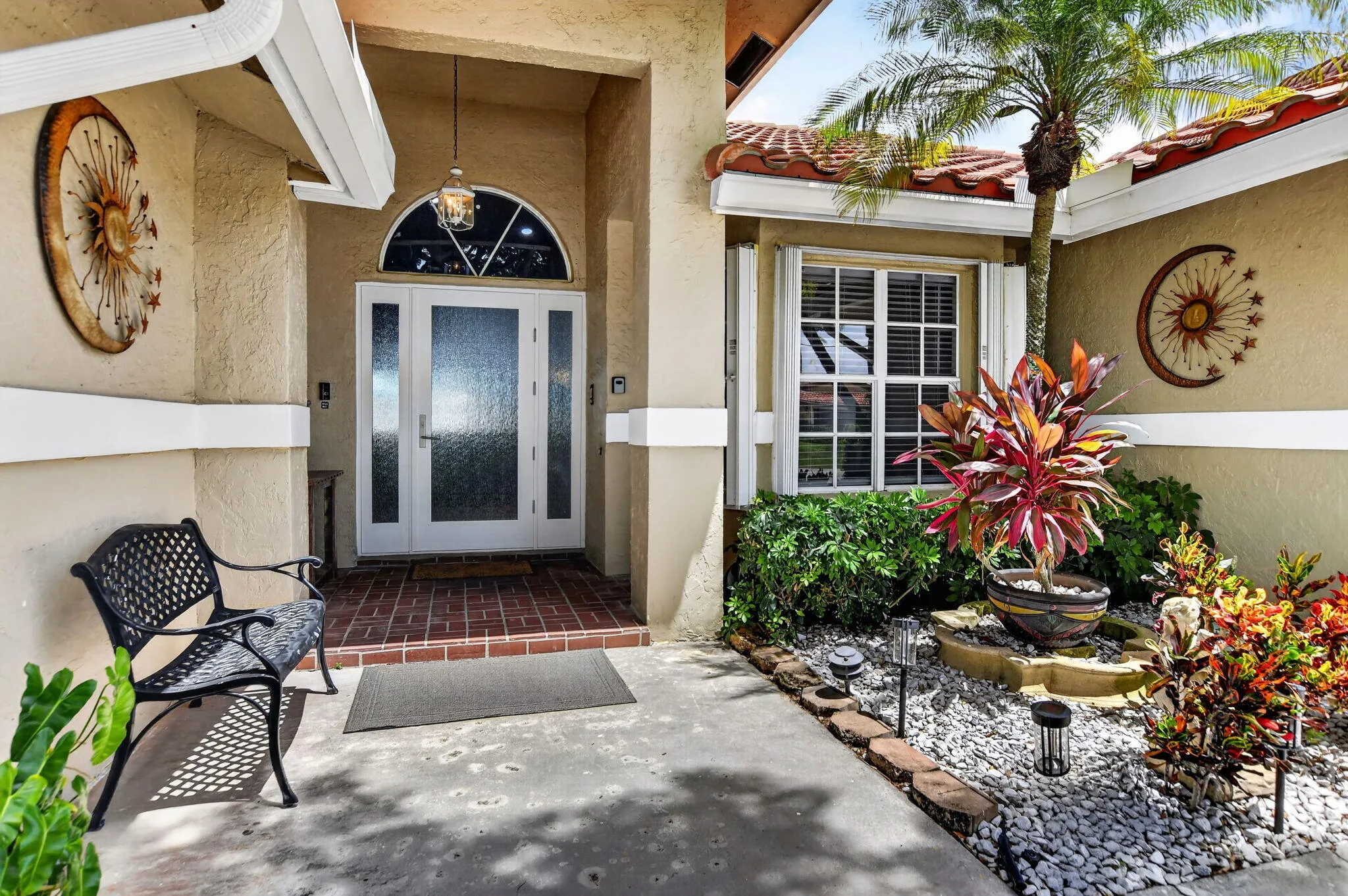 Arched transom window above double entry doors with frosted glass panels, brick-finish flooring at entry, window with white blinds, mature palm tree, and decorative landscaping with white pebbles