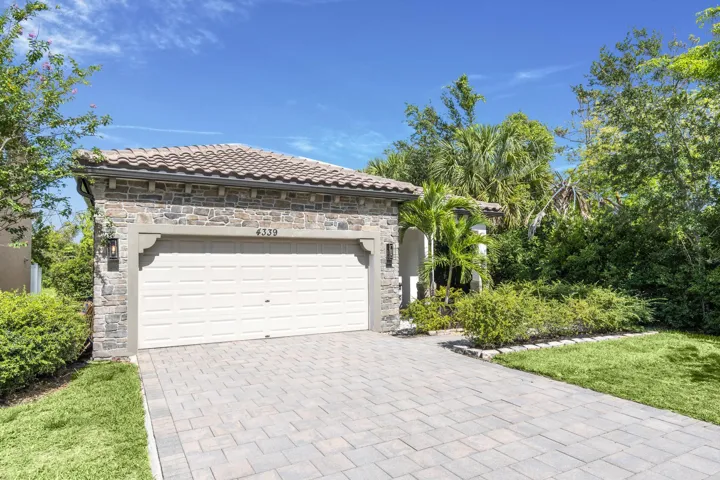 Modern residence featuring a pristine white garage door with clean-lined paneling, wide paver driveway in neutral tones, lush mature landscaping with tropical and deciduous trees, manicured lawn, and a tile roof with contemporary architectural detailing