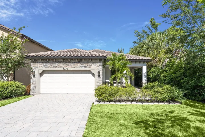 Stucco exterior with stone veneer accents, tile roof, paver driveway, attached garage, and mature landscaping