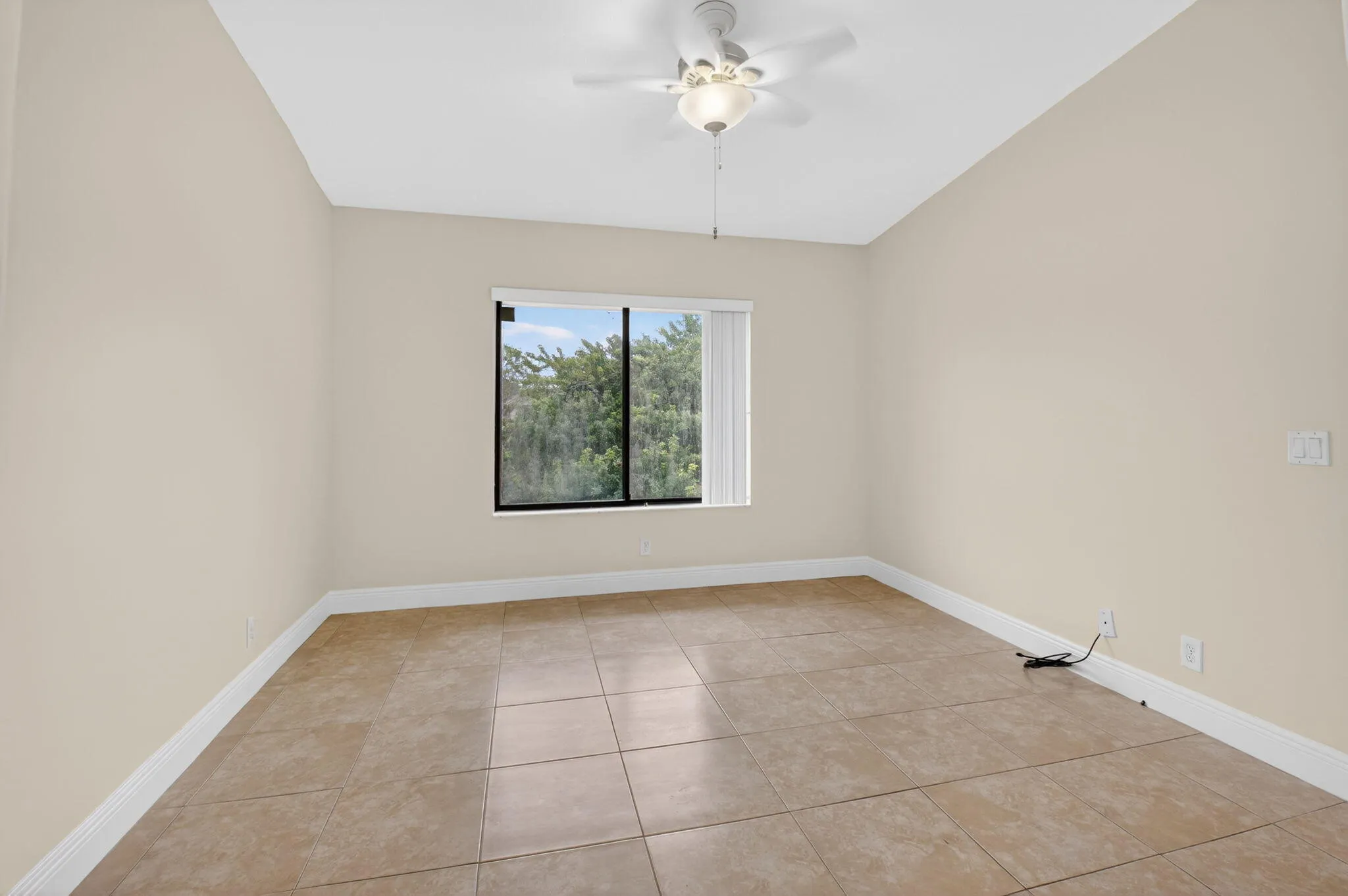 Spacious room featuring neutral-toned walls, tiled flooring, a window offering exterior scenery, and a ceiling fan with integrated lighting