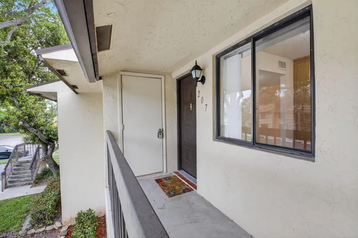 Exterior balcony with a dark railing, leading to a dark entry door with an attached exterior light fixture and a white side door