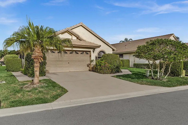 Concrete driveway, attached garage, well-maintained landscaping with mature shrubs and a palm tree, and a tiled roof