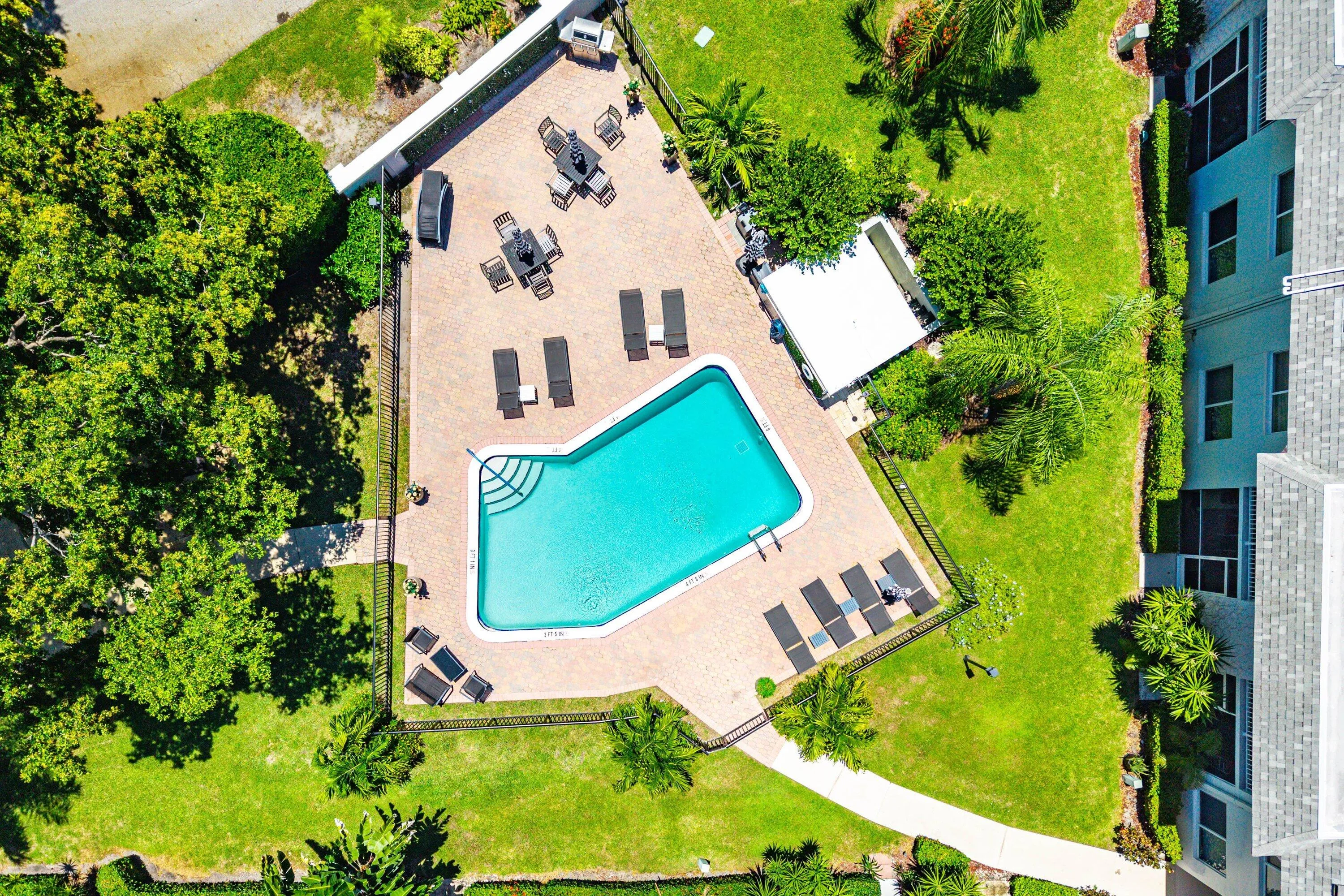 Paved patio with an in-ground swimming pool, surrounded by green landscaping, and a fenced perimeter