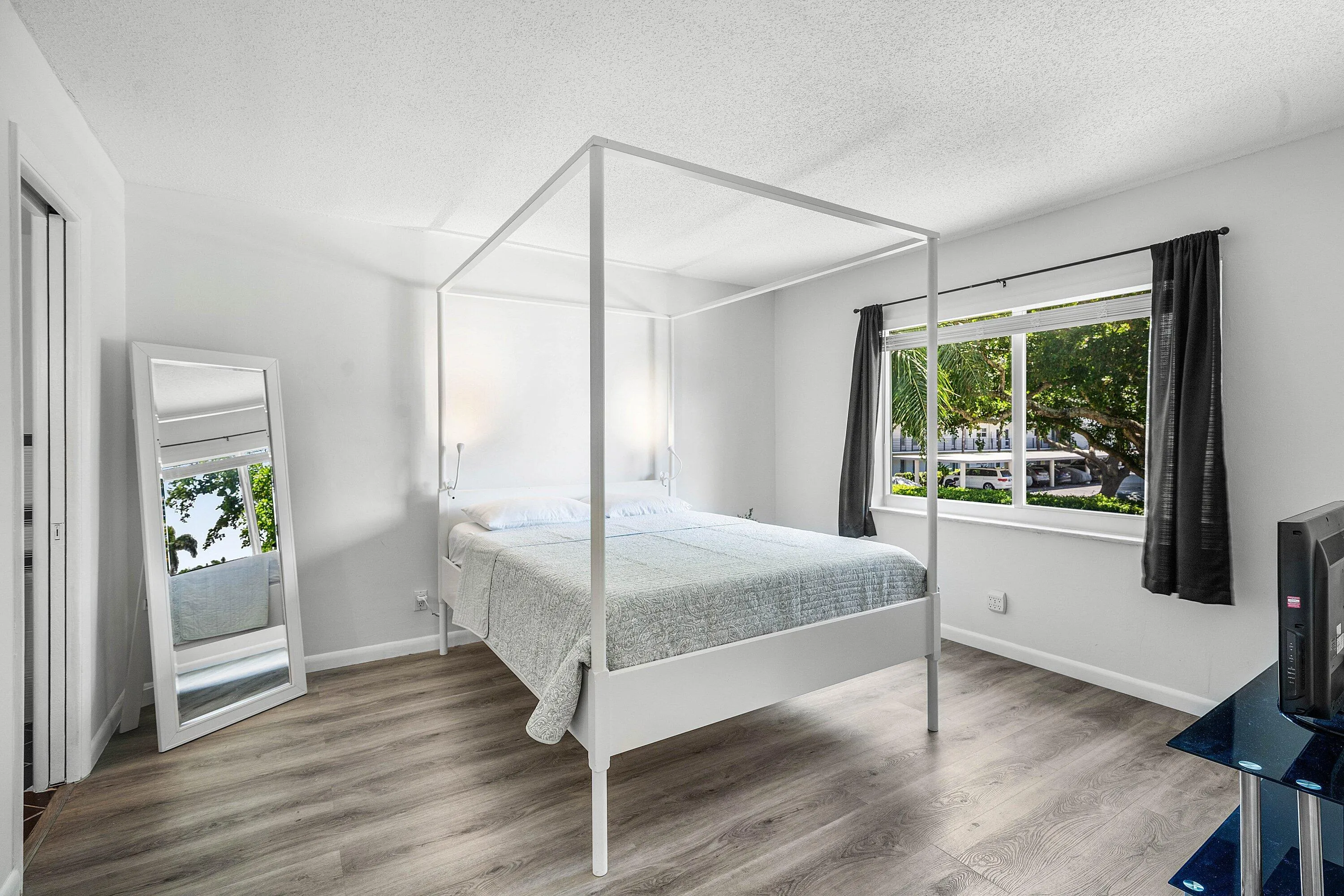 Bright primary bedroom featuring a four-poster canopy bed frame, wood-finish flooring, and large windows with natural light