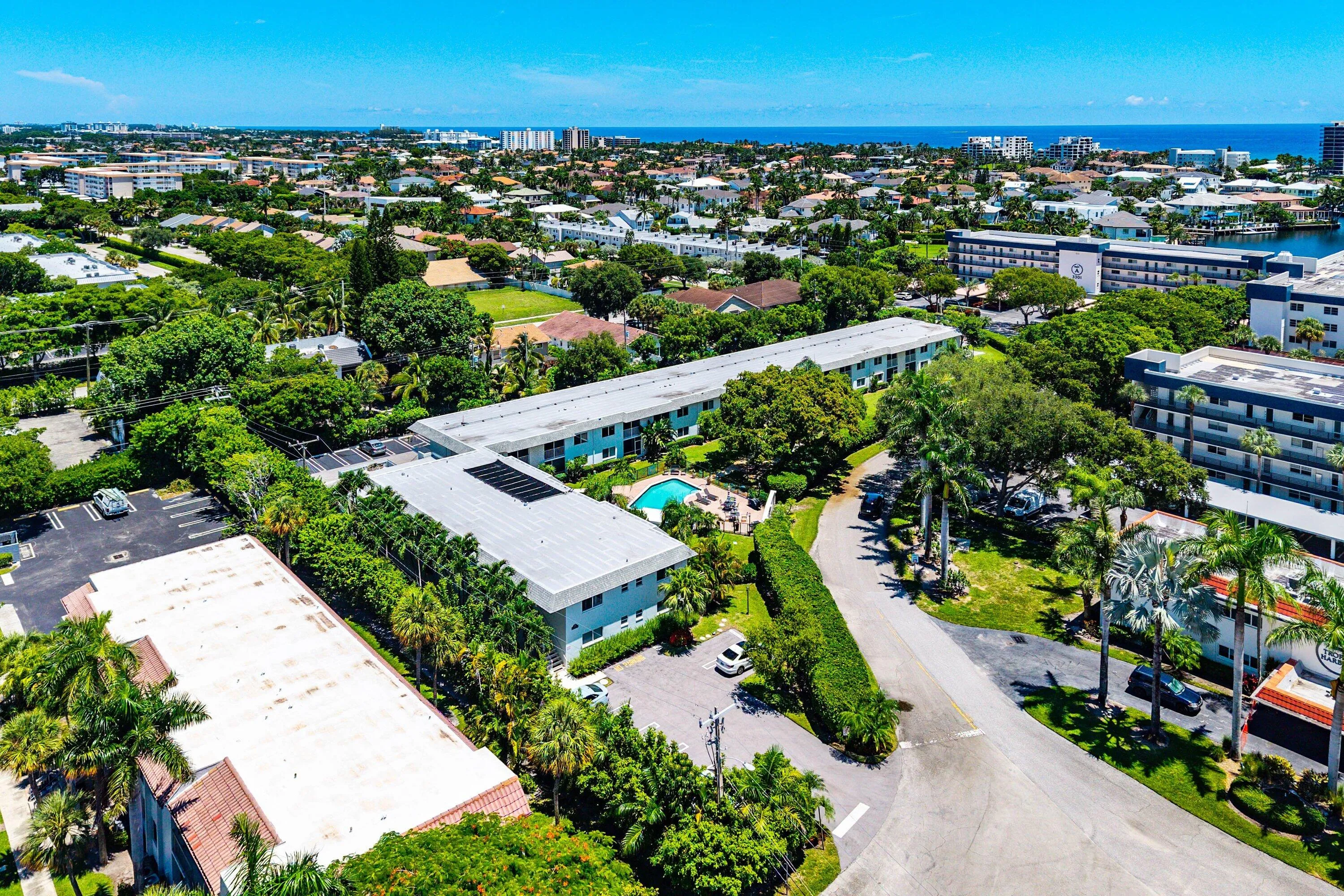 Aerial perspective showcasing multiple buildings, lush landscaping, a swimming pool, and proximity to a body of water and distant cityscape