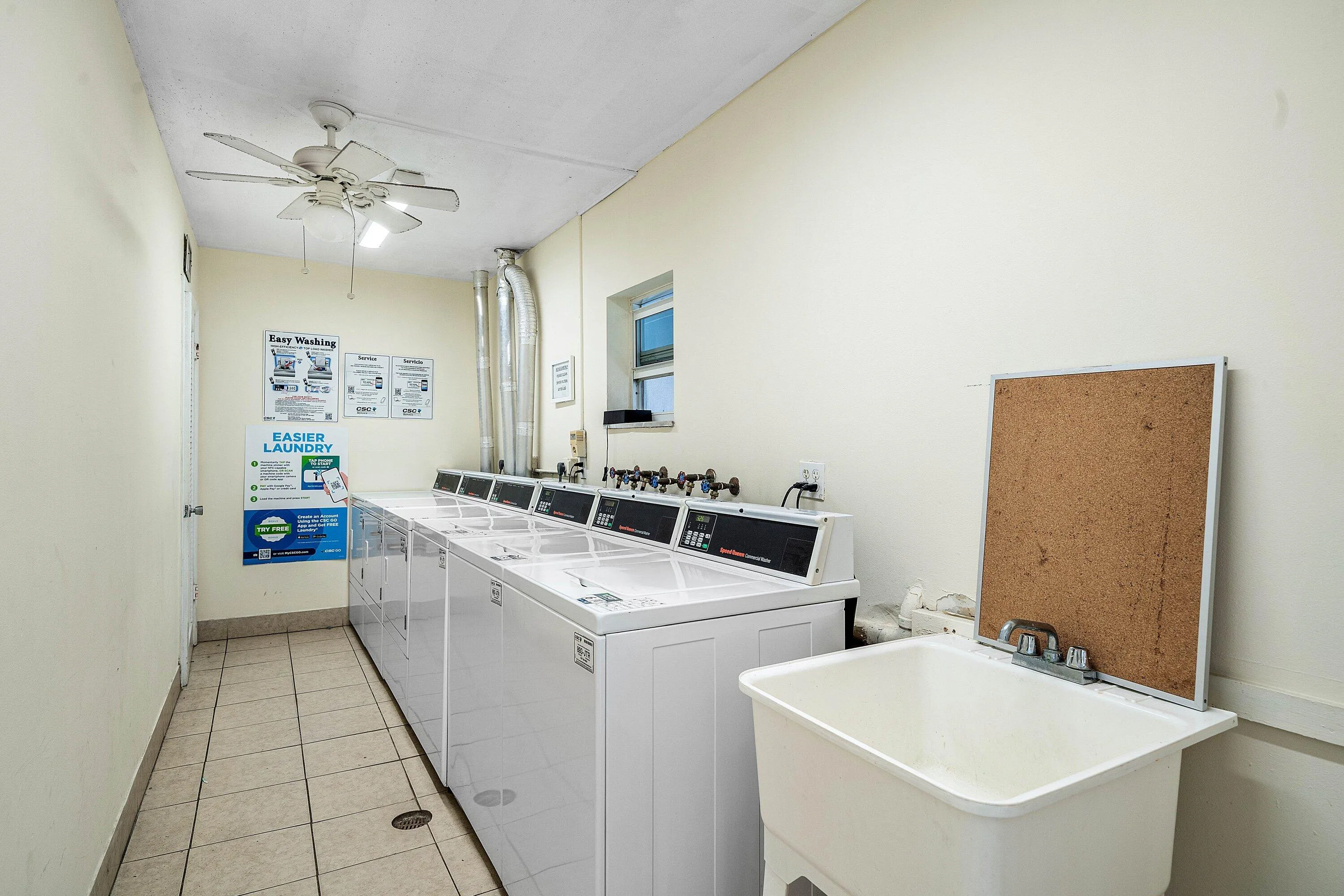 Multiple white top-load washing machines, utility sink with chrome faucet, tiled flooring, and a ceiling fan
