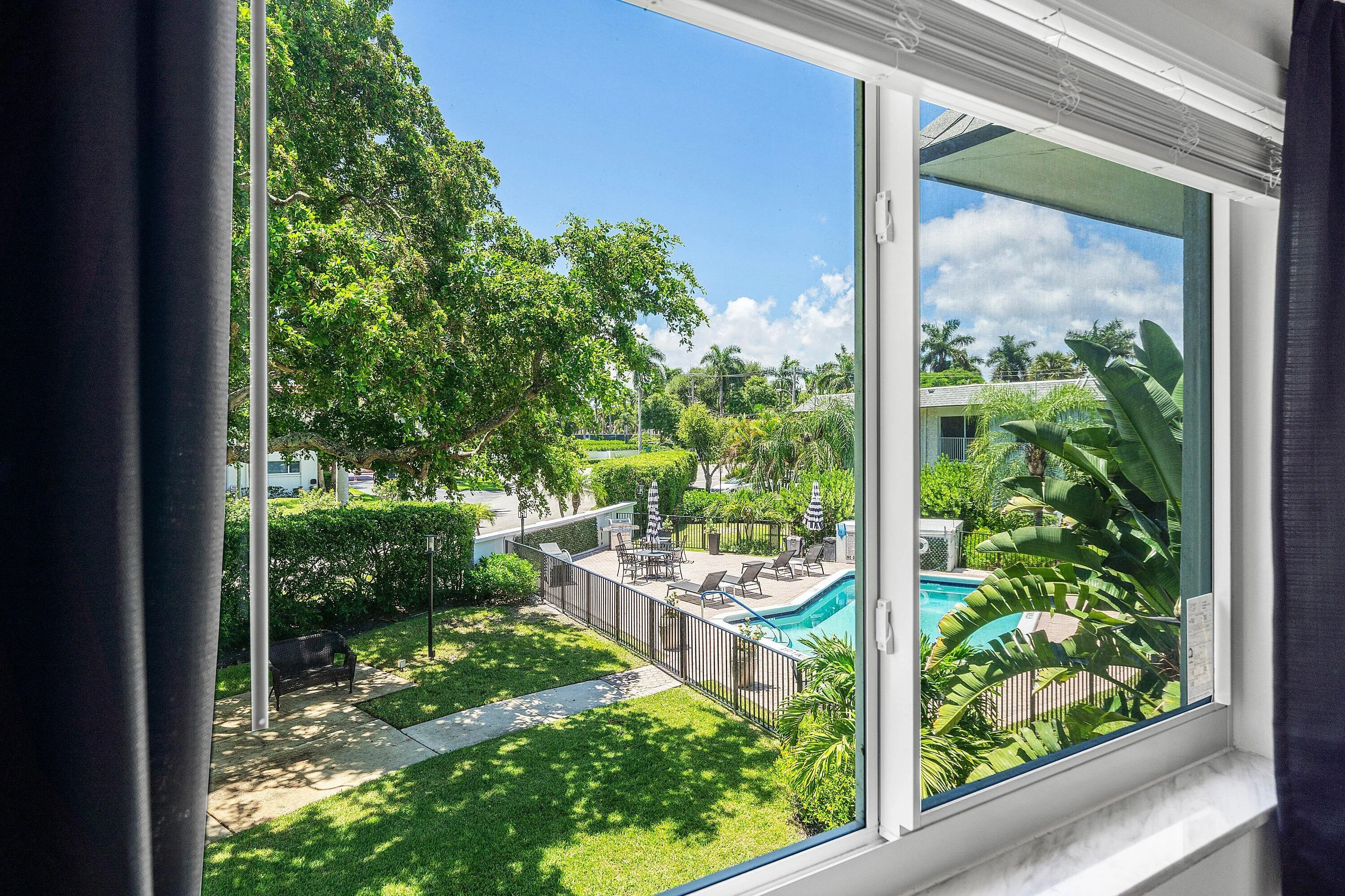Bright, light-filled interior framed by expansive glass doors opening to manicured grounds featuring mature landscaping, lush lawn, and resort-style amenities including an in-ground swimming pool and spa area with hardscaped decking